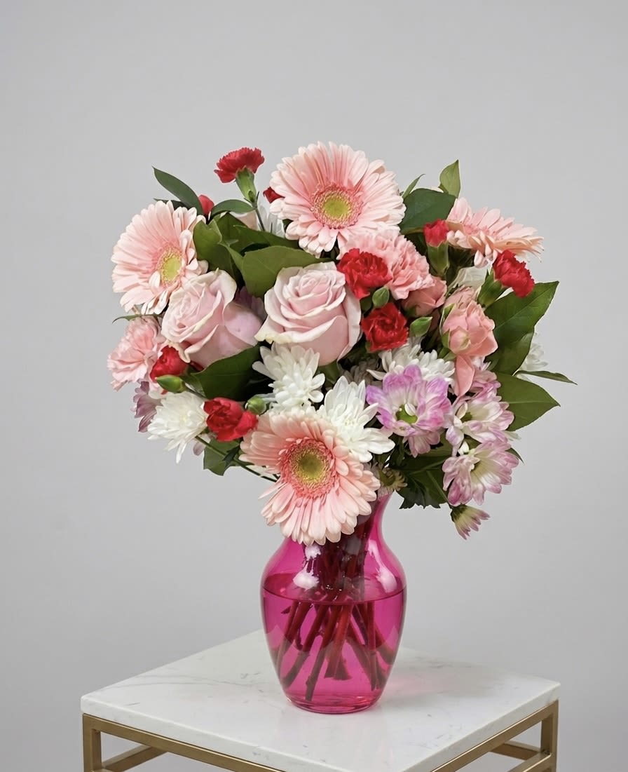 The Blushing Luxe - Elevate your space with "The Blushing Luxe," a professionally designed, mixed-flower arrangement that marries bold color with elegant textures. This striking centerpiece features perfectly formed pink Gerbera daisies—the stars of the show—with no missing petals, creating a full and uniform look. They are artfully layered with rich, deep-hot pink  carnations, delicate light-pink carnations, and pristine white chrysanthemums, all set among lush green foliage.