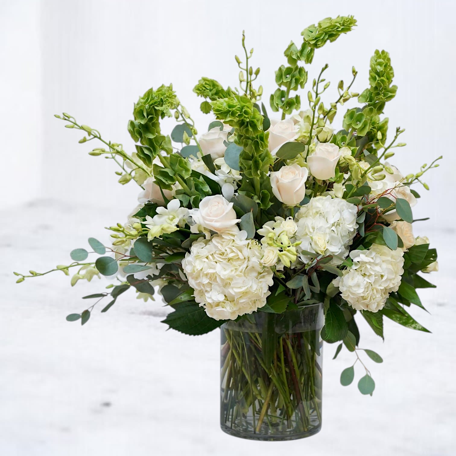 Sunday Morning - Sunday Morning is a lush white and green flower arrangement designed with layered white hydrangea, premium roses, fresh orchids, bells of Ireland, and eucalyptus in a modern glass vase. Filled with soft texture, airy movement, and fresh tonal greens, this arrangement brings together an elevated garden-style aesthetic with a clean editorial feel.  Designed for sympathy flowers, luxury everyday gifting, birthdays, thank you flowers, housewarming gifts, celebrations of life, and elegant home styling, Sunday Morning blends timeless white blooms with sculptural greenery for a naturally refined look that feels both calming and sophisticated.  Handcrafted with premium seasonal flowers and available for same-day flower delivery in Magnolia, TX and surrounding areas including The Woodlands, Montgomery, Conroe, Spring, Tomball, and Lake Conroe communities.