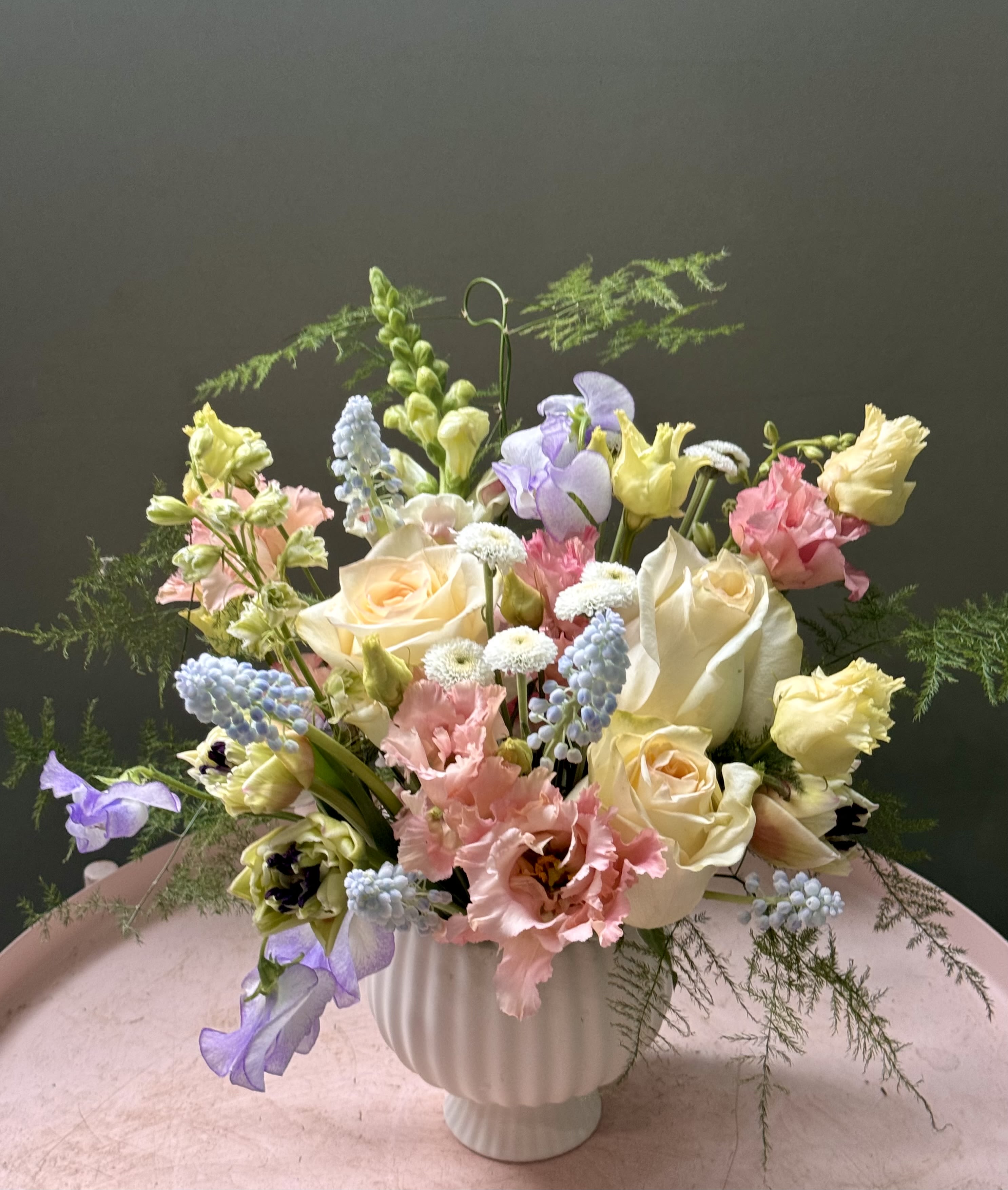 Delicate  - Soft pastels and a garden-fresh feel make this birthday bouquet or Mother's Day arrangement feel like a gentle embrace. The Delicate arrangement features cream and white roses and soft pink lisianthus up front, with lavender snapdragons and pale yellow blooms weaving through airy, feathery greenery-perfect for anniversaries, spring celebrations, or a thoughtful just-because gift for someone who loves romantic, understated florals.  Clusters of light-blue muscari/hyacinth-like accents and classic daisy shapes add sweet texture while the loose, rounded silhouette keeps the design feeling natural and unstuffed. Arranged in a white fluted ceramic vase, the piece stands with an elegant, timeless profile-she'll feel genuinely seen when this arrives on her table. Order as shown for a ready-to-display size that arrives hydrated and arranged; simple care: fresh water and a cool spot will keep the blooms looking their best.