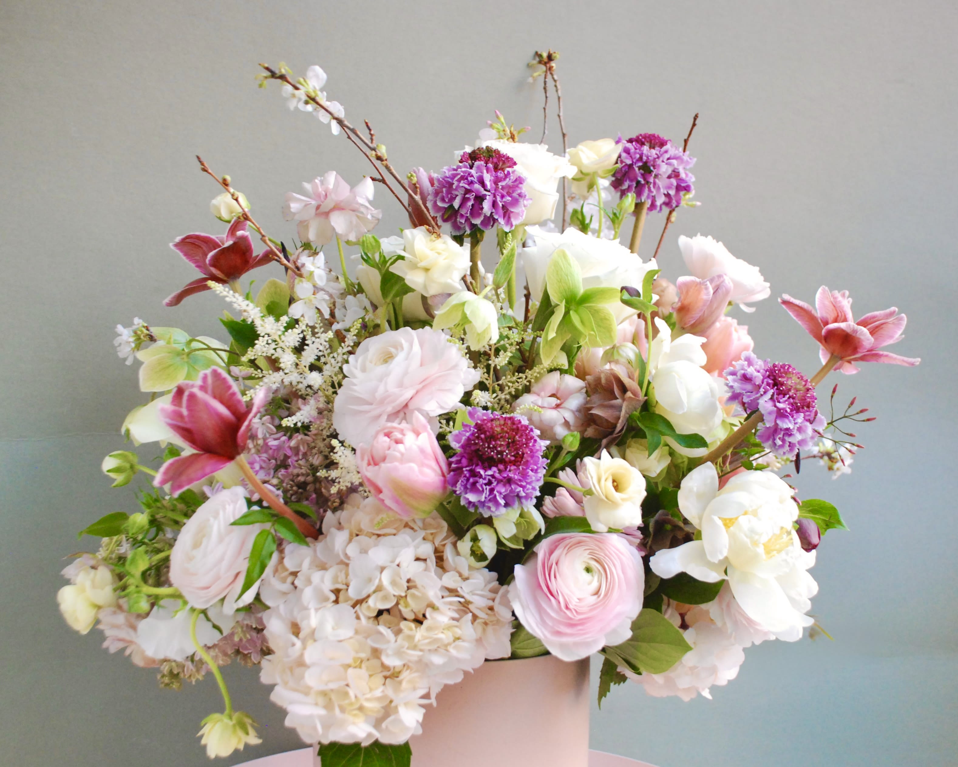The one  - A soft pastel garden bouquet in blush pinks, creamy whites, lavender and spring green makes a tender statement for a wedding bouquet, anniversary roses gift, or a thoughtful springtime birthday bouquet. The arrangement reads airy and romantic-the kind of floral surprise that will make the person you're thinking of feel truly seen and celebrated without being over the top.  Rounded hydrangea clusters and layered ranunculus mingle with rose-like blooms, tulip-shaped flowers, delicate dianthus and lilac accents for a lively mix of textures and sizes. Branching stems add height and movement while smaller clustered flowers fill the composition, all gathered in a pale ceramic vase that complements the soft color story.  Perfect for brides, honored guests, or anyone who loves a garden-fresh look, this arrangement arrives loose and abundant for an effortless, celebratory feel. It's offered in a large, airy style that works well on a dining table, entryway, or as a graceful gift; change the water every few days and trim stems slightly for best vase life.