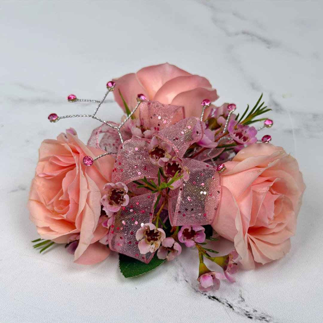 Rose Quartz 3 Rose Wrist Corsage - A cluster of blush pink roses accented with tiny pink filler blooms creates a soft, romantic focal point for this Rose Quartz 3 Rose Corsage, perfect for prom night and other special occasions. This corsage reads elegantly feminine at first glance: the small, rounded composition sits neatly on the wrist and catches the eye without overpowering any outfit.  Crafted with three spray roses and matching filler blooms in pink, this compact arrangement is finished with a sheer pink glitter ribbon threaded through the center for gentle shimmer. Pink decorative rhinestone gem picks with thin silver wire accents add subtle sparkle and formal polish, making the piece feel celebratory and refined. Attached with a white lace wristlet, it's easy to attach and designed to stay secure through an evening of photos and dancing; she'll look and feel truly celebrated.