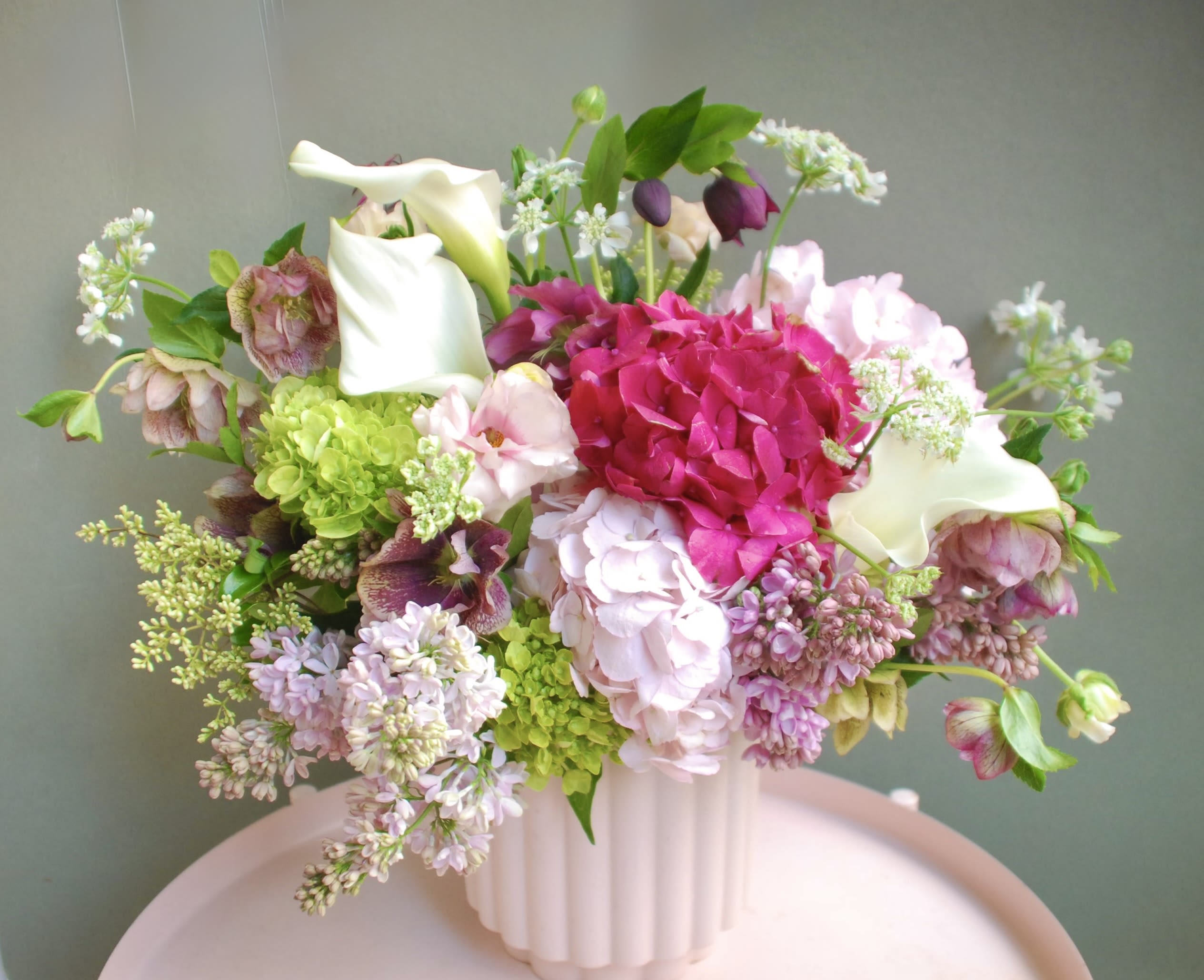 Mamma Mia  - A show-stopping hot-pink hydrangea cluster anchors this Mamma Mia bouquet, creating an immediate burst of color that feels both romantic and joyful-perfect as a birthday bouquet or a springtime gift. The arrangement reads as an anniversary or celebratory floral gift that will make the person you're thinking of feel cherished the moment it arrives.  Soft pale-pink hydrangeas and crisp white calla lilies surround the vivid center, while lavender and mauve lilac tones, small clustered filler blooms, and airy branching accents give the design a loose, garden-style silhouette. Textures range from the fluffy, voluminous hydrangeas to the sleek calla lilies, creating contrast and movement across a fresh pink, white, green, and purple palette. The arrangement comes in an elegant ribbed ceramic vase that complements its natural, effortless look and makes it ready to display.  Ideal for moms, partners, or a friend celebrating a milestone, this arrangement sends a message of warmth and thoughtful celebration without being overformal. Arrange care is simple-keep the vase filled with fresh water and trim stems every few days to extend the life and keep the flowers looking full and vibrant.
