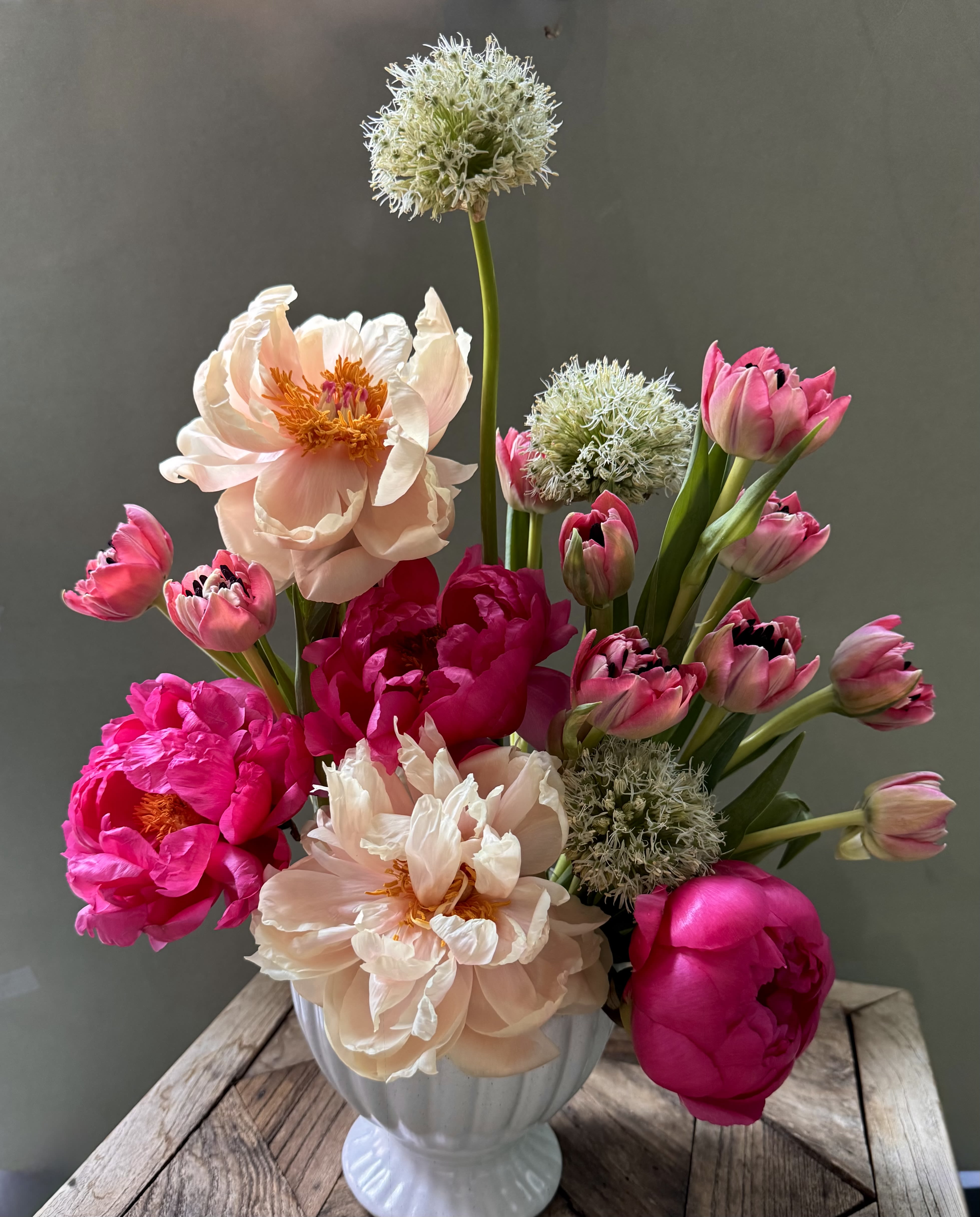Maria  - Large blush and hot-pink peonies steal the show in this spring-ready flower arrangement, paired with pink tulips and round white allium-like blooms for a fresh, romantic look perfect as a birthday bouquet or celebratory centerpiece. The upright, garden-style composition gives the recipient a sense of joyful abundance-she'll feel seen and celebrated by the lively mix of soft pastels and saturated magenta accents.  Arranged in a clean white ceramic vase, Maria combines full, layered peony blossoms with strong vertical tulip stems and spherical white blooms to create contrast in texture and shape. The palette of pink, white, and green reads light and elegant from every angle; the vase's neutral finish makes it right at home on a mantel, dining table, or office desk.  Ideal for spring occasions, anniversaries, or simply to say "I'm thinking of you," this bouquet sends a warm, uplifting message without being over the top. For best enjoyment, keep the water fresh and the arrangement out of direct sunlight to extend the life of the peonies and tulips.