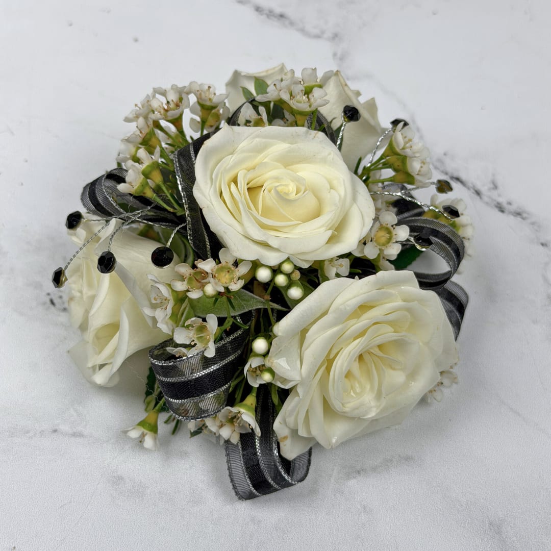 Onyx 4 Rose Wrist Corsage - Cream-white roses contrasted with black and silver shimmering ribbon make this wrist corsage a quietly dramatic pick for prom or any formal evening event. The Onyx 4 Rose Corsage blends the classic romance of roses with modern contrast so the person you're celebrating looks polished and unforgettable on the dance floor.  Four cream-white roses are nestled among tiny white filler blossoms for soft texture and a fresh, natural look. Black and silver ribbon loops and decorative gem-like rhinestones create an artful, tailored finish, all held together on a white lace wristlet for easy wearing. The restrained color scheme reads elegant and adaptable, perfect for an outfit that deserves some striking contrast.