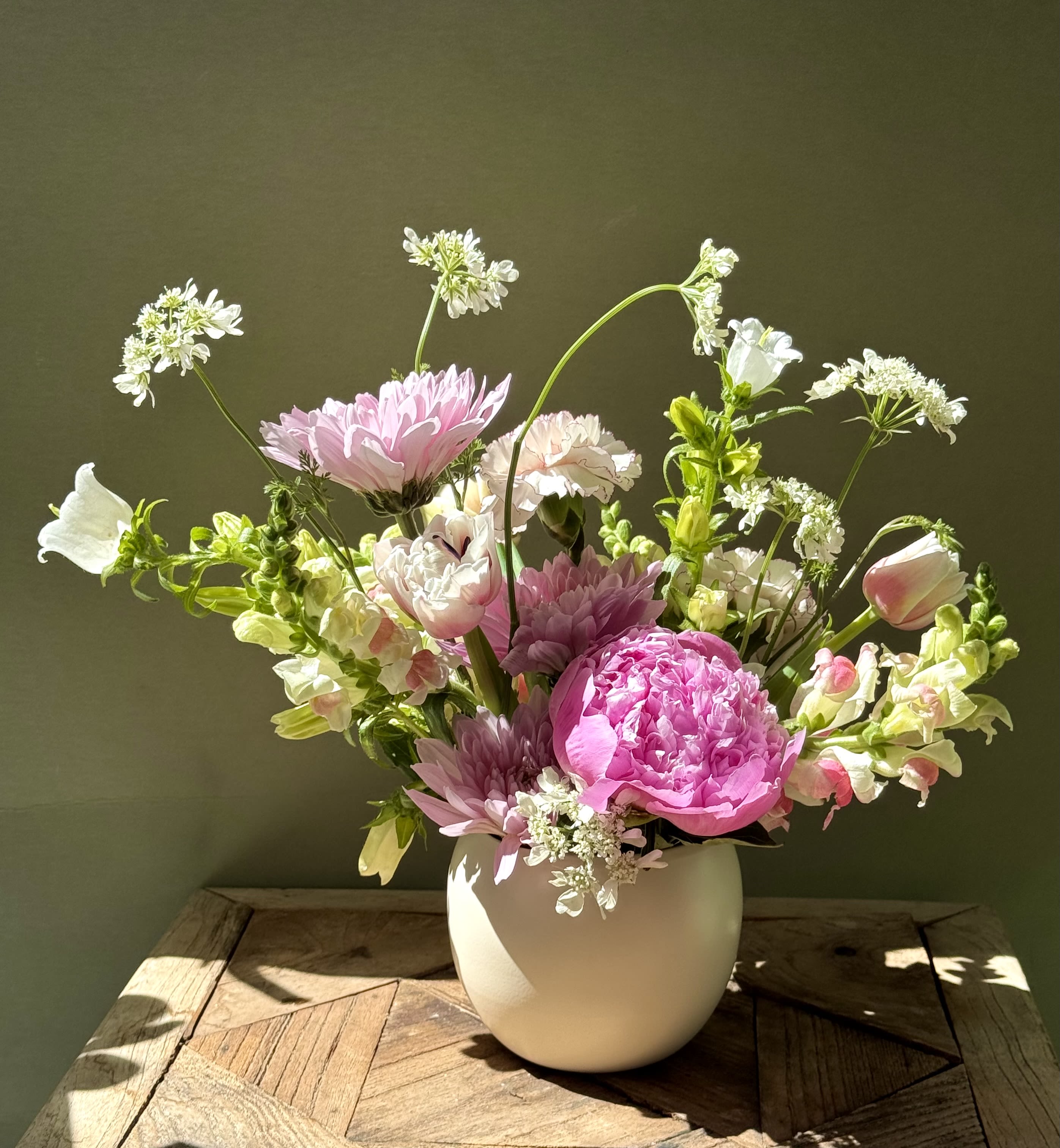 Mom’s Garden  - A soft, garden-style arrangement that reads like a spring bouquet in a white ceramic vase - perfect as a birthday bouquet or a gentle way to say "thinking of you." The rounded, open shape places a large bright pink, peony-like rose at the center, surrounded by pale pink and white daisies and tulip-shaped blooms that give a fresh, feminine feel. This floral arrangement makes the recipient feel cherished and celebrated with its romantic, light-filled palette.  Delicate snapdragon spikes and airy white filler flowers add vertical movement and a relaxed, natural silhouette, while ruffled carnations bring texture and fullness. Displayed in a simple white ceramic vase, the composition measures as a rounded, outward-arching design that suits spring gifting, everyday celebrations, or a thoughtful get-well gesture. Arrange on a table or bedside to brighten a room; fresh-cut stems benefit from daily water checks to keep the mix looking its best.