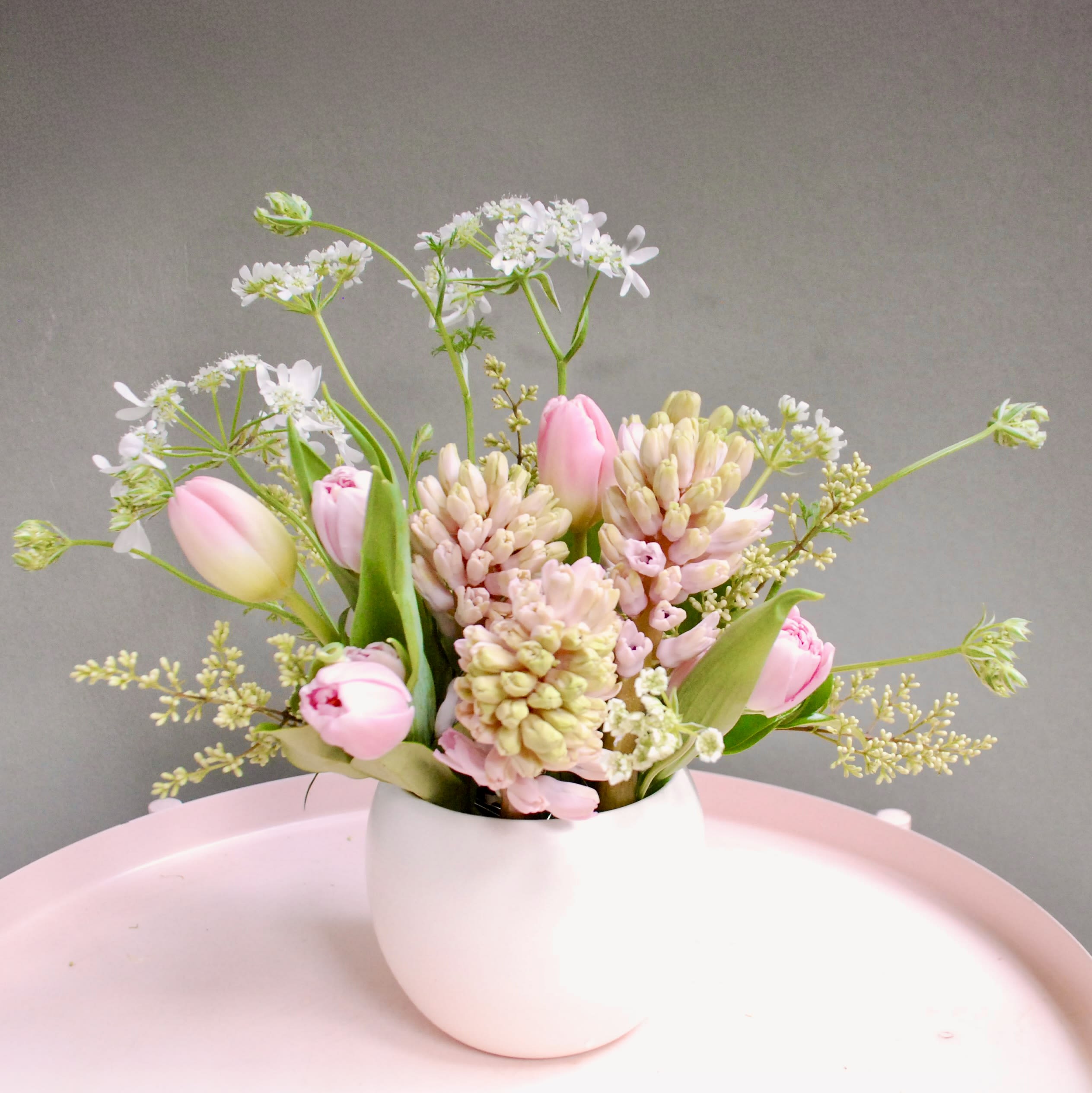 Julia - Soft pink tulips and clusters of pale pink and cream hyacinth-like blooms mingle with airy white filler in a loose, garden-style arrangement-perfect as a springtime birthday bouquet or a Mother's Day gift. The rounded white ceramic vase and asymmetrical, open structure give the display a light, romantic feel that will make the person you're thinking of feel seen and celebrated without being overdone.  The palette blends pink, white, green, and touches of soft yellow for a fresh, pastel look. Stems are arranged loosely to create a gentle, textured silhouette that works equally well as a bedside surprise, a cheerful thank-you, or a soft decorative centerpiece. The white ceramic vase keeps the focus on the blooms and fits easily into most home decor.