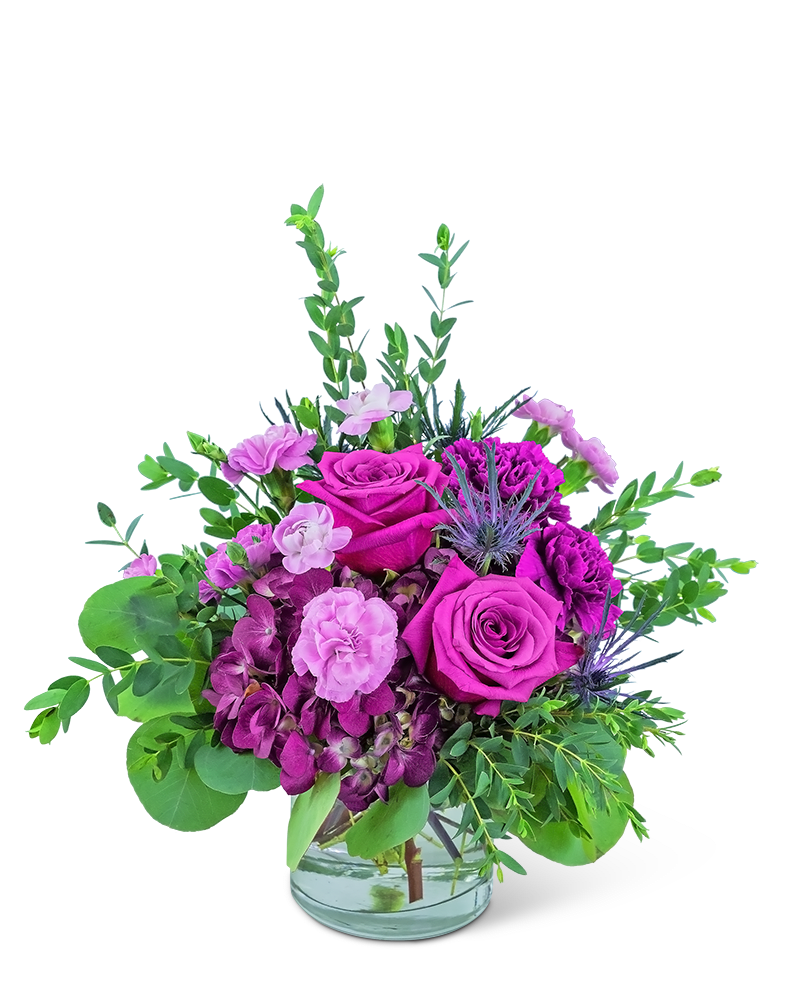 Low purple and pink rose arrangement in a clear glass vase with accent blooms