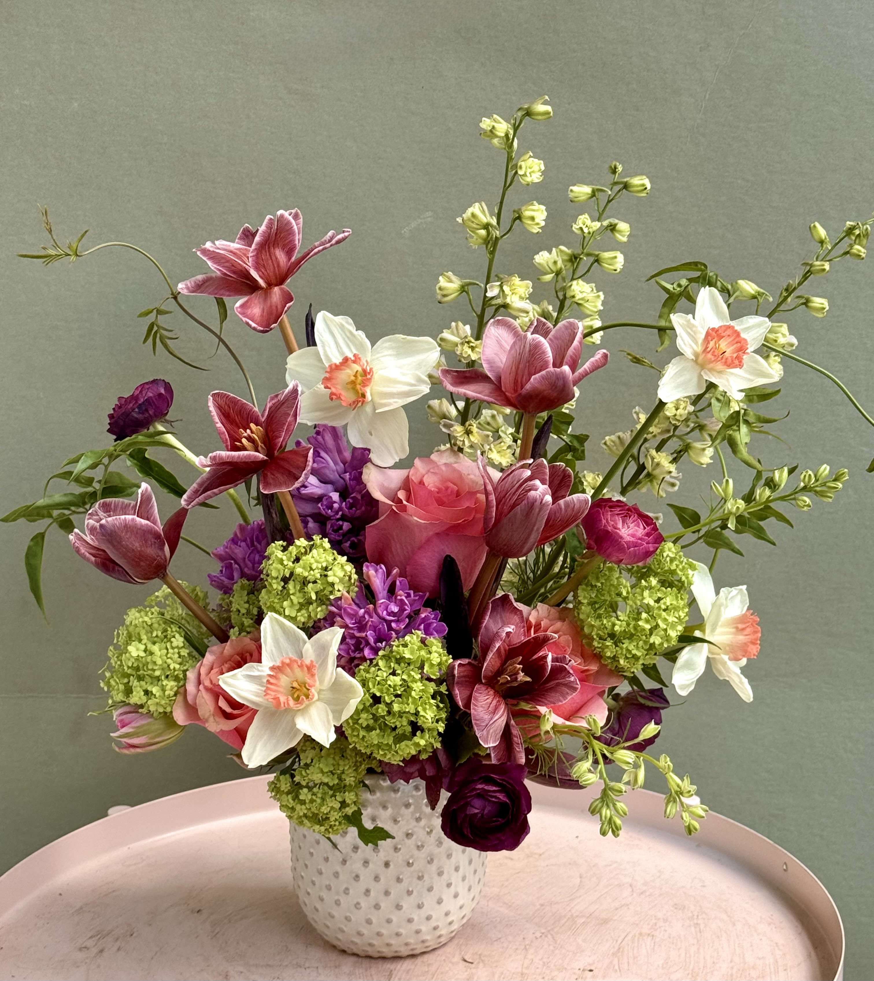 Magali  - Pink tulips and purple hyacinth form the first impression of this springtime flower arrangement, perfect as a birthday bouquet or a cheerful "thinking of you" gift. The tall, sculptural silhouette makes it an elegant anniversary roses alternative or a stunning centerpiece for a seasonal celebration, sending a fresh, romantic message to the person you're honoring.  This arrangement features pink tulips, white daffodils, purple hyacinth and garden-style roses arranged loosely with bright green clustered filler for volume and contrast. The long stems give an airy, sculptural look while the mix of soft pastels and saturated purples creates lively visual texture. Flowers arrive in a white textured vase set on a pale pink tray for a polished presentation that's ready to display.  Ideal for spring occasions or anytime you want to brighten someone's day, Magali suits recipients who appreciate romantic color and graceful height. Standard care: keep water fresh, recut stems every few days, and place away from direct heat to extend vase life.