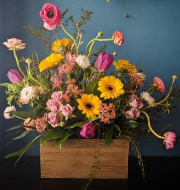 Rustic Spring Serenade -  Rustic Spring Serenade  This whimsical garden-style arrangement is a joyful celebration of spring’s playful beauty. Designed in a simple natural wooden box, it features many of Spring's best blooms: cheerful golden gerbera daisies dancing among delicate pink ranunculus, lavender tulips, blush spray roses, and soft white accent blooms. Airy textural greenery and trailing botanical elements create movement and an organic, free-flowing silhouette. Each design will vary due to availability and seasonal products.   The asymmetrical design, Flemish Style, gives it a fresh-from-the-garden feel, while the vibrant contrast of sunny yellows and romantic pinks brings warmth and charm. With its loose, artistic composition and rustic container, this arrangement feels both modern and naturally elegant—perfect for brightening a space with effortless springtime sophistication.