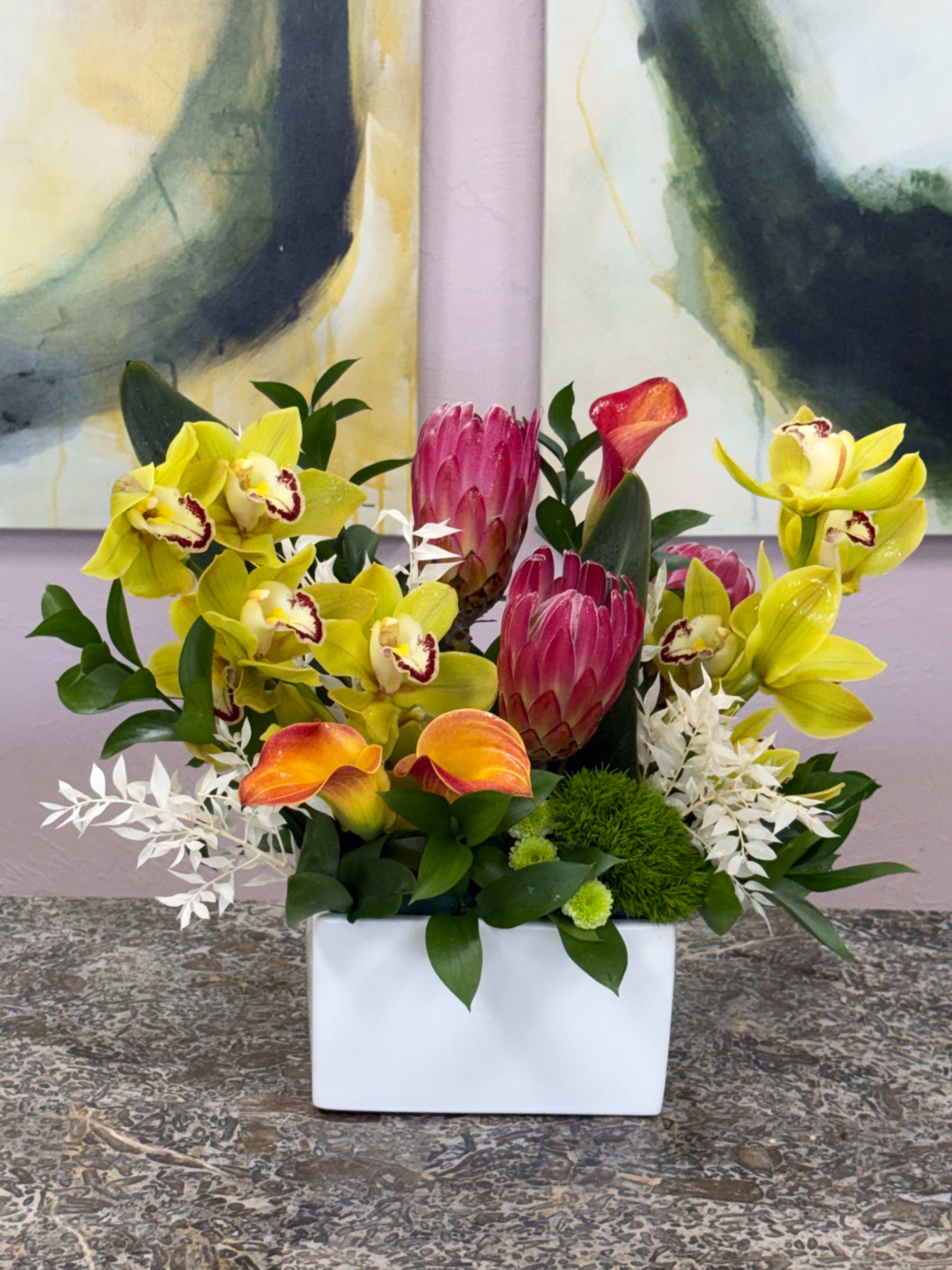 Sun-woven Citrus - This dense, layered composition pairs orange calla lilies with small white accent blooms and glossy dark foliage for high-contrast drama. Textured green pom-pom accents add playful depth, and the flowers are arranged in a low white rectangular ceramic vase that suits tabletops, mantels, or office desks. The palette of yellow, pink, orange, white, and green creates a cheerful statement that stays stylish and chic.  For gifting, this arrangement is the kind of bouquet that makes the person you're thinking of feel truly seen-warm, vibrant, and thoughtfully modern. Its compact size fits intimate spaces and requires minimal care: keep flowers in fresh water and out of direct heat to extend bloom life. Order for same-week delivery to ensure the freshest presentation for birthdays, new homes, or everyday celebrations.  *Flowers are subject to substitutions of equal value.