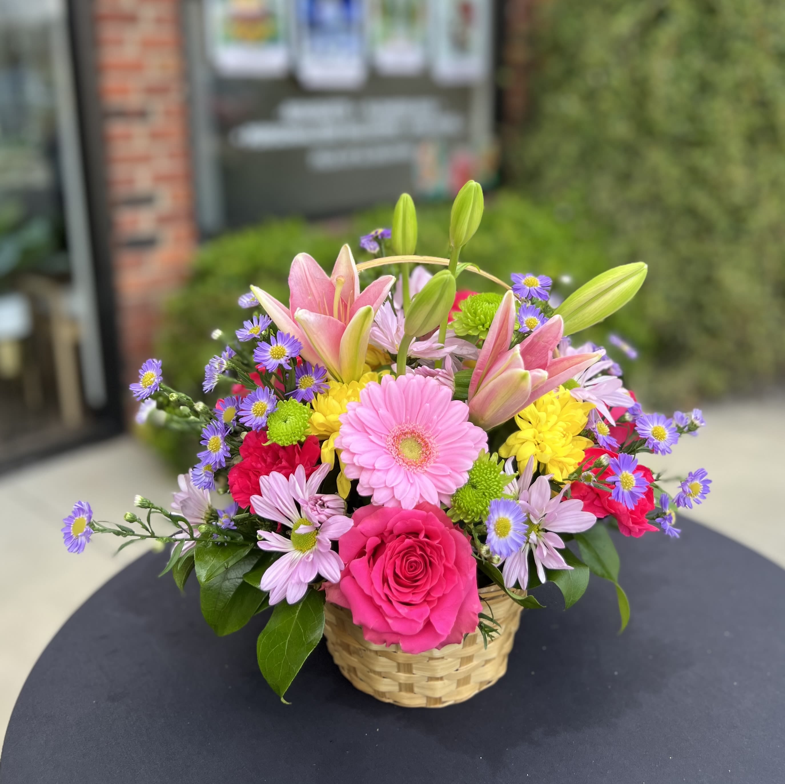 Daisy  - Sunny, garden-fresh color greets the recipient at first glance - Daysi is a cheerful flower arrangement perfect for a birthday bouquet, a bright "thank you," or a just-because pick-me-up. The lively mix of pink lilies and a vivid pink rose creates a warm focal point while lavender and sunny yellow daisy-like blooms lift the composition; small green button mums add playful texture so the person you're thinking of will feel cheerful and noticed.  Arranged low in a woven basket, this casual, approachable design reads like a handful of spring from the market: saturated pinks, soft purples, and sunny yellows woven together with green accents. The compact shape makes it easy to set on a table or desk without overwhelming the space. Perfect for someone who prefers an informal, upbeat gift, Daysi arrives ready to display - simply place in a cool spot and refresh the water as needed to help blooms last.