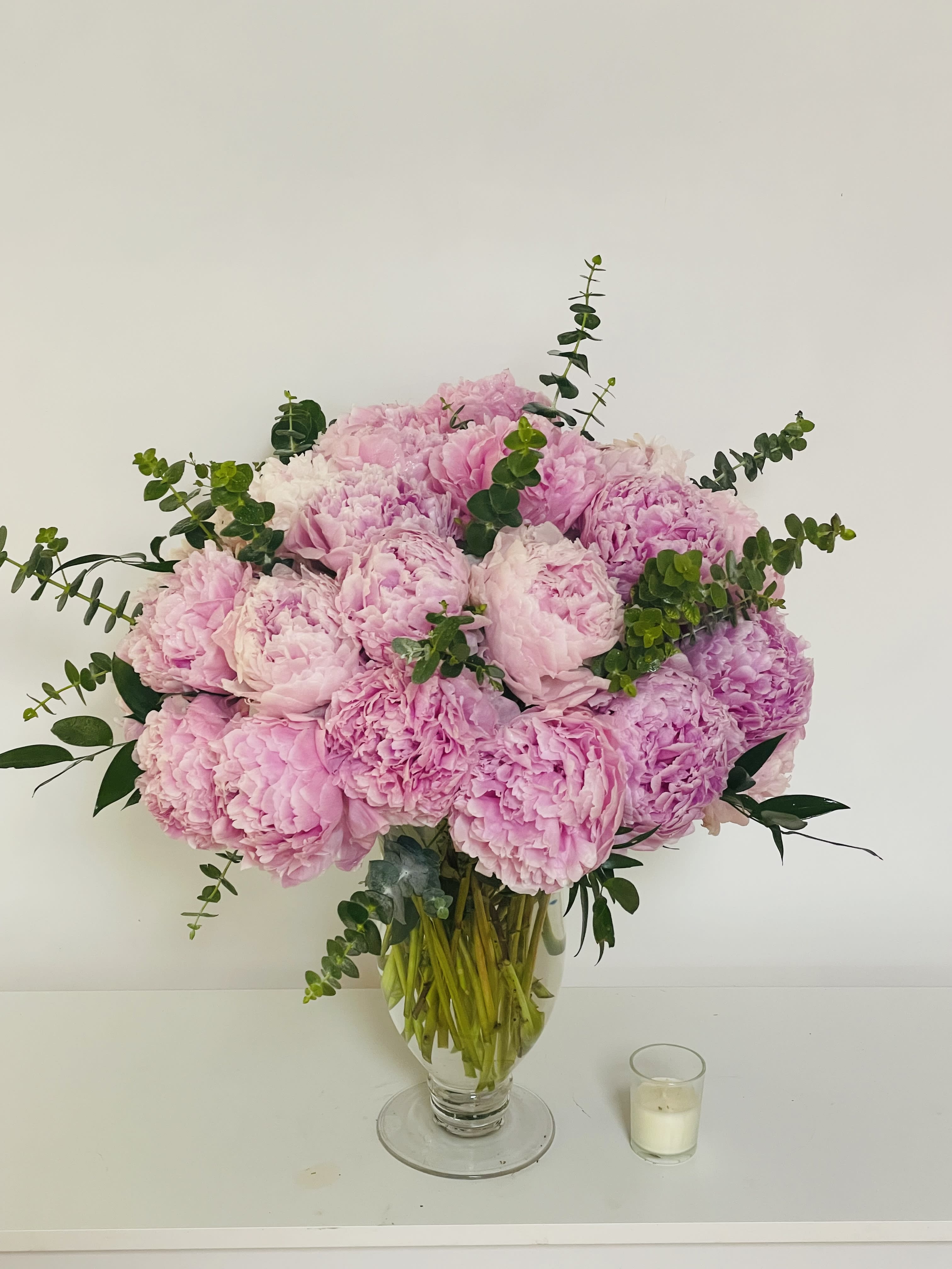 Large arrangement of pink peonies in a clear glass vase beside a small votive candle