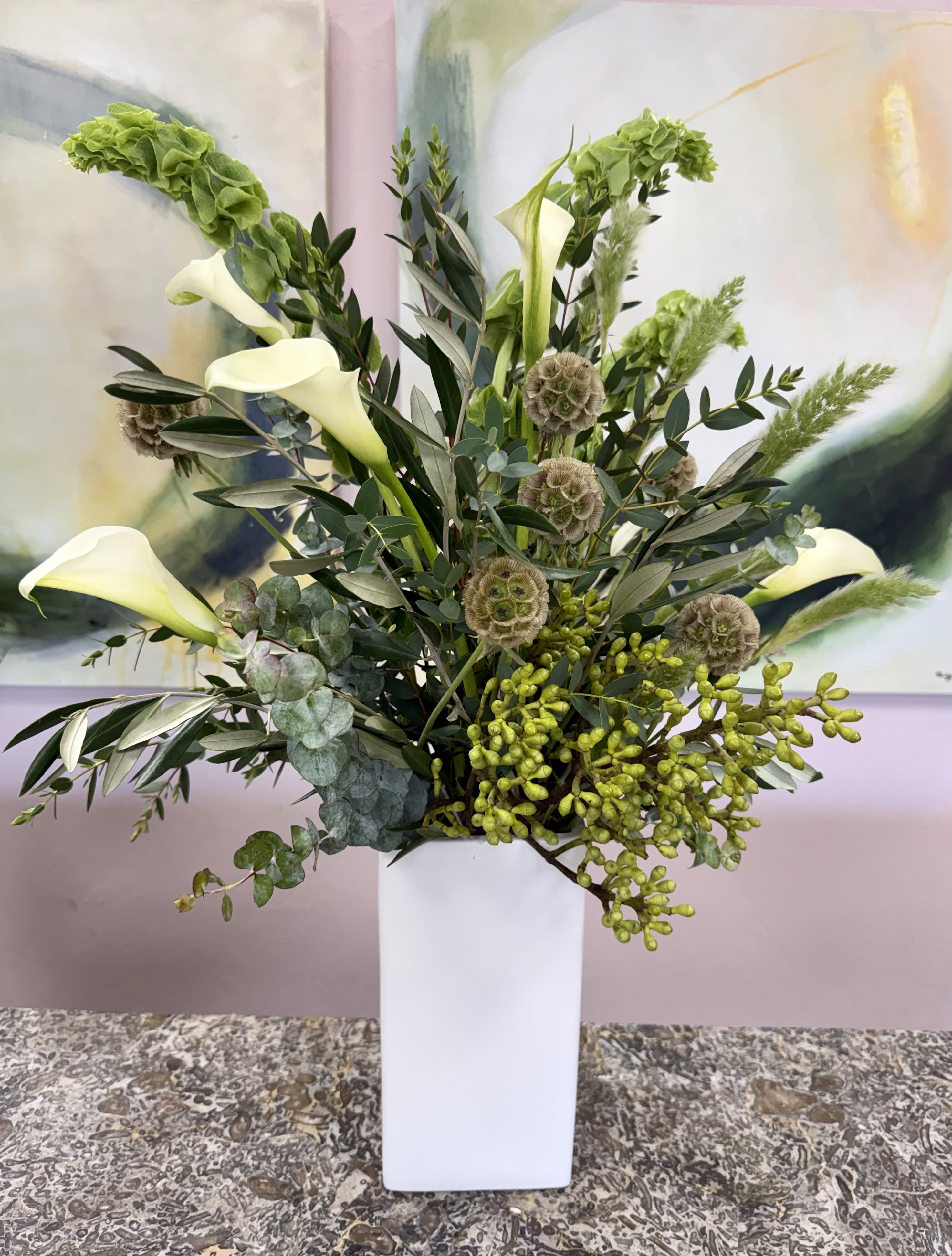 Naples Tide in Green - Tall white calla lilies rise like sculpted candles through a textured sea of greenery-this flower arrangement is a modern, serene choice for a thoughtful present or elegant everyday display. As a flower arrangement designed to soothe and impress, Naples Tide in Green presents a fresh green and white palette that reads calm and refined, the perfect way to let someone know you're thinking of them or to elevate a reception table.  The design is built in a tall white rectangular ceramic vase and pairs several mini calla lilies with a loose, airy mix of foliage. Broad glossy leaves provide sculptural contrast while rounded eucalyptus-like sprigs and clustered green accents add layered texture and gentle movement. The overall effect is light and contemporary, with clean lines and a soft, natural scent from the lilies and greenery.  Made to sit tall and make a quiet statement, this arrangement suits bedside tables, entry consoles, altars, or narrow dining runners-anywhere the recipient should feel comforted or celebrated without overpowering the room. If you'd like adjustments for size or to include a message card for delivery, select your options at checkout so the flowers arrive personalized for the moment.  *Flowers are subject to substitutions of equal value.