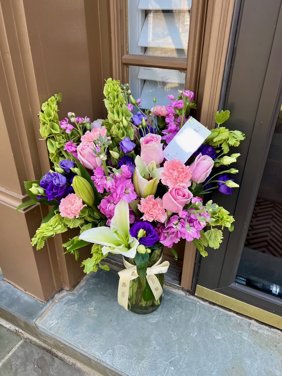 Rosita  - Large, lush bouquet of pink roses, white lilies, pink carnations ,  pink stock , purple lisianthus and greenery in a glass vase with ribbon.