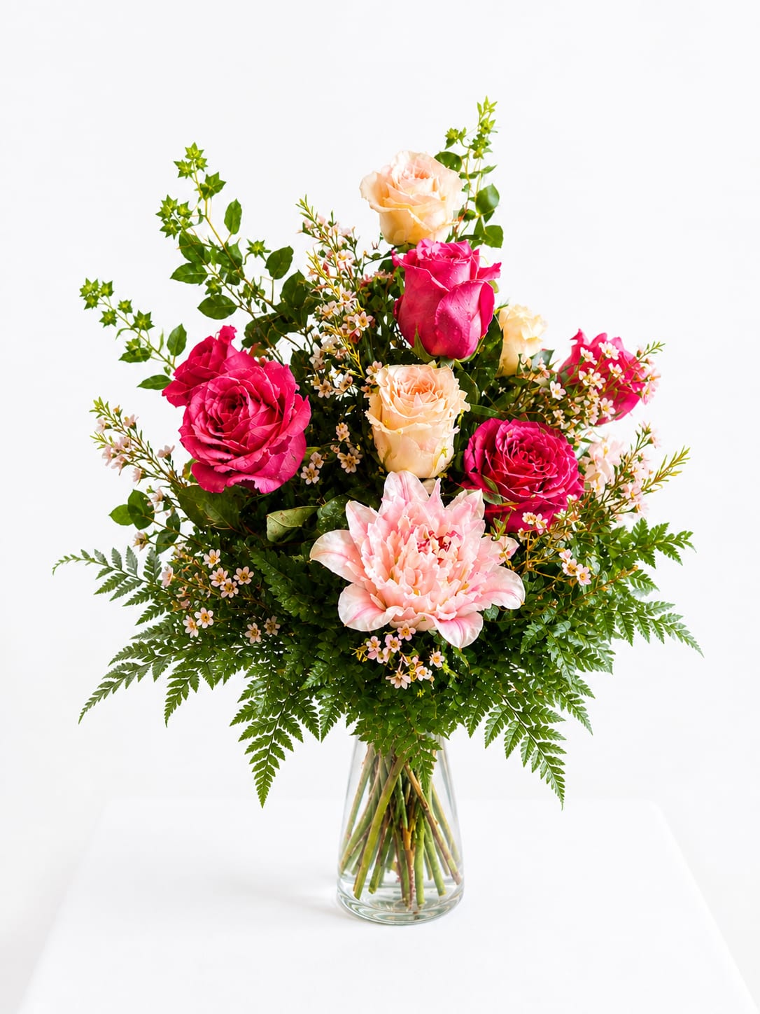 Lace &amp; Love Blooms (10% off Mother's Day Special) - Lush clusters of hot pink and peach roses surround a large pale-pink lily to create a romantic, garden-style flower arrangement perfect for a birthday bouquet, anniversary flowers, or a warm thinking-of-you gift. The bright pinks, soft blush, and touches of yellow and white give the arrangement an instantly cheerful, celebratory mood that will make the person you're thinking of feel truly seen and cherished.  This full, airy design mixes roses, lilies, and delicate filler blooms with long, branching greenery and glossy leaves for a layered, just-picked look. Unopened lily buds add height and movement while tiny white and pale-pink fillers soften the composition; stems are presented in a clear glass vase tied with a sheer white ribbon for a polished, classic finish. Ideal for everyday gifting, Mother's Day, or a thank-you gesture - simple care: keep water fresh, recut stems every few days, and enjoy the vase-worthy display for several days to come.