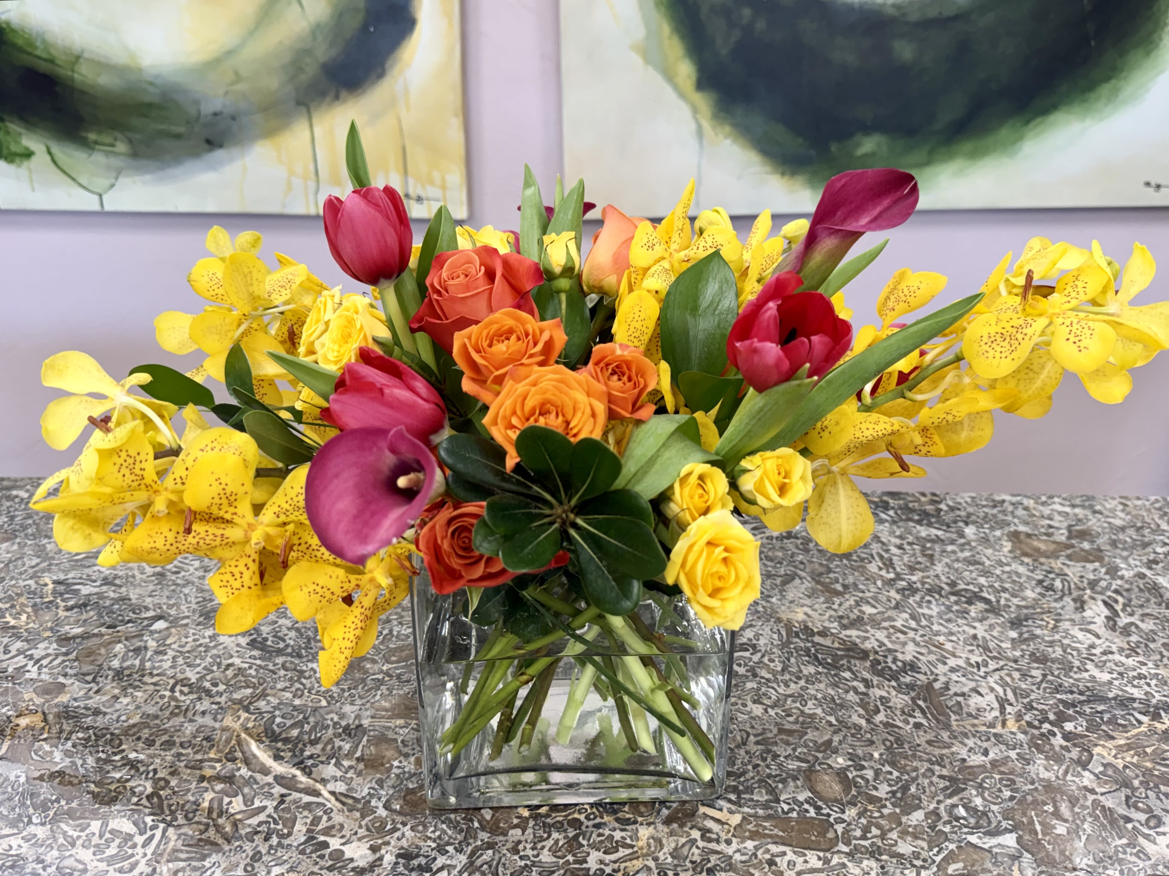Naples Sunset Riot - Bright color pops make this flower arrangement a joyful choice for exciting gift. The Naples Sunset Riot arrangement pairs vivid yellow orchids with orange-and-yellow roses and touches of magenta blooms , to create a high‑contrast, celebratory look that makes the recipient feel energized and truly noticed.  Arranged low and wide in a clear glass vase for a contemporary presentation. The composition is lush and abundant rather than delicate-saturated yellows and oranges warm the eye while the purple calla lilies add dramatic depth. Perfect for someone who loves bold color and modern style.  This arrangement suits birthdays, housewarmings, congratulations, or any moment you want to brighten-she'll feel instantly uplifted when it arrives. The low vase works well on dining tables or coffee tables; keep flowers in fresh water and out of direct heat to maintain peak color and longevity.  *Flowers are subject to substitutions of equal value.
