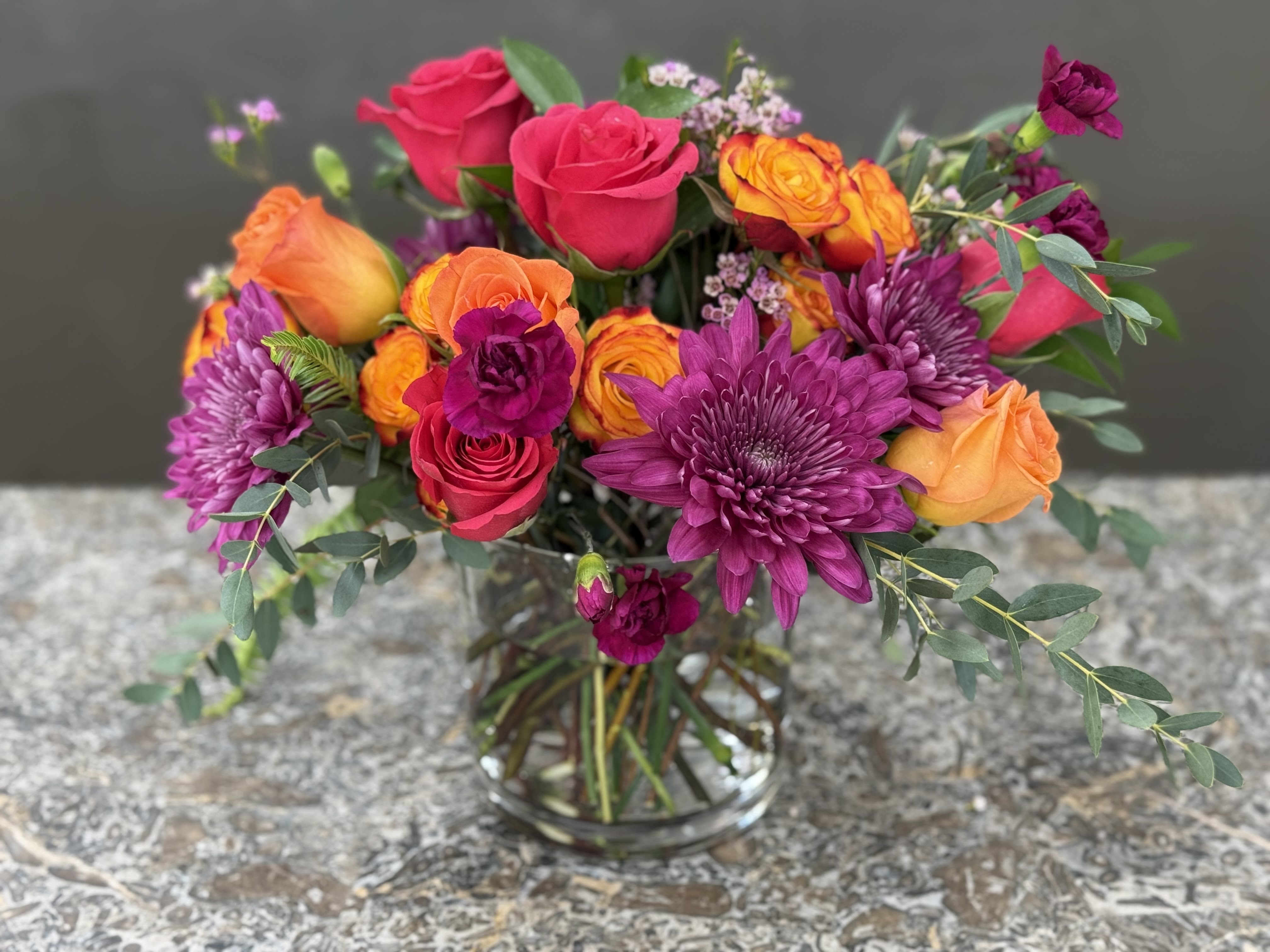 Sweat Heat - A lush cluster of hot pink, orange and purple blossoms greet the eye - a cheerful everyday bouquet that reads romantic and celebratory at once. This mixed-rose arrangement, styled for birthdays, thank-yous, or just-because moments, blends saturated sunset colors create a lively impression the recipient will notice the moment it arrives.  This is the kind of bouquet that makes someone feel celebrated and seen, whether it's for a birthday, a bright thank-you, or a spontaneous romantic gesture. Vase-ready and low-maintenance, replace the water every few days and trim stems for longer life; sized to sit on a table or desk, it arrives ready to brighten a room.  *Flowers are subject to substitutions of equal value.