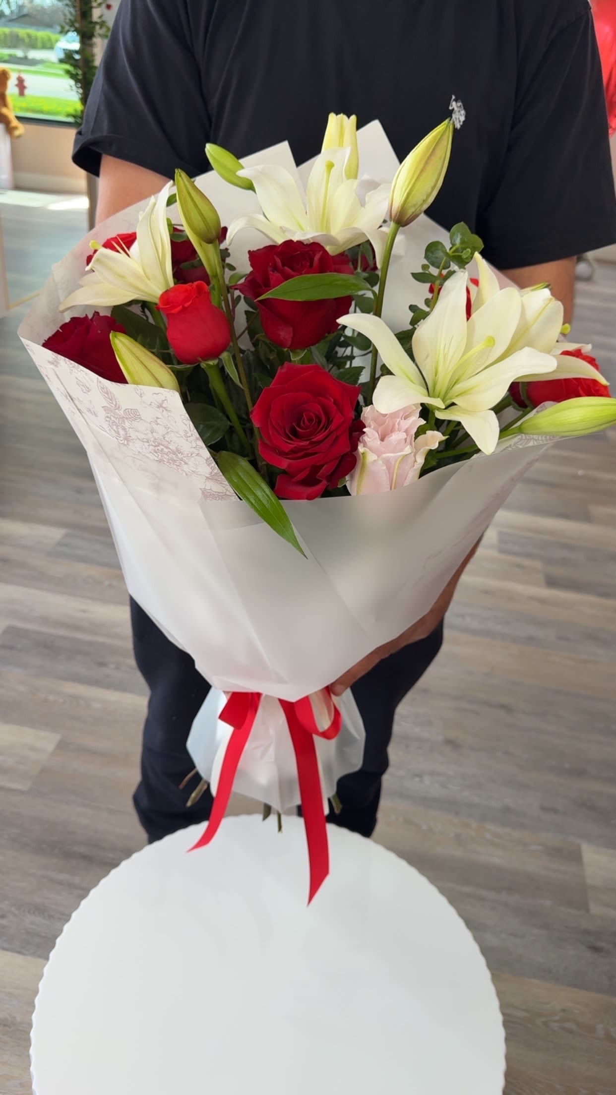 Love in Bloom - Deep red roses and large white lilies come together in a romantic flower arrangement made for anniversaries, Valentine's gestures, or any time you want to say "I love you" with classic elegance. The strong red-and-white contrast is softened by a pale pink carnation tucked among the blooms, so the recipient will feel both noticed and cherished the moment they see it.  The bouquet features multiple deep red roses, open white lilies, and a small pale pink carnation arranged tightly for a lush, textured look. One presentation is wrapped in crisp white paper and tied with a red ribbon for a formal, hand-held bouquet; another option arrives arranged inside a white round gift box for an extra-polished reveal. The mix of rose buds and open lilies creates visual depth and a fresh fragrance profile.  Perfect for anniversaries, romantic surprises, or a meaningful thank-you, this arrangement reads as thoughtful and refined-ideal for the person you want to make feel truly seen. Choose the wrapped bouquet or the gift-box presentation when ordering; avoid direct sunlight and refresh the water regularly for the longest vase life.
