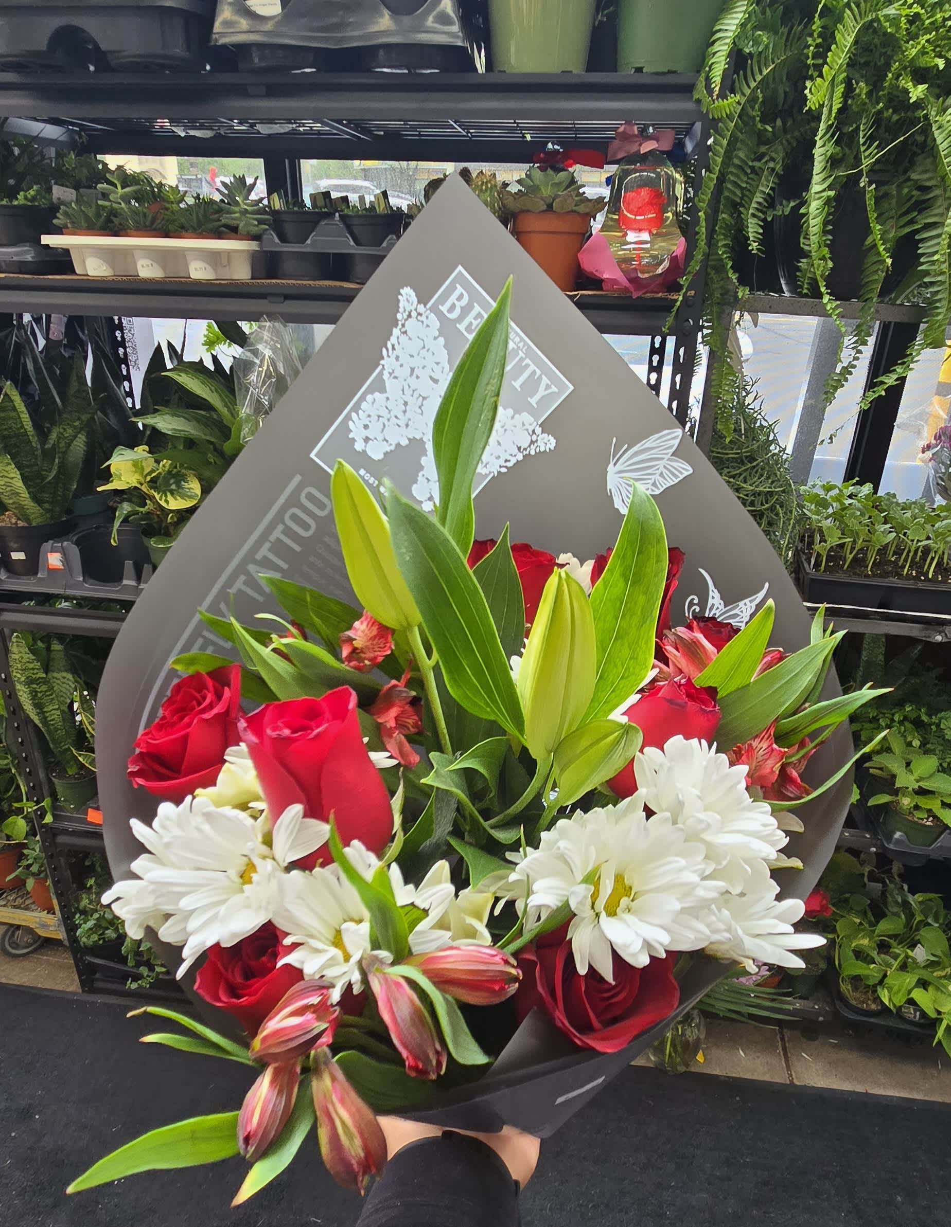 Handheld bouquet of red roses, white daisies, lilies, and alstroemeria in gray paper wrap