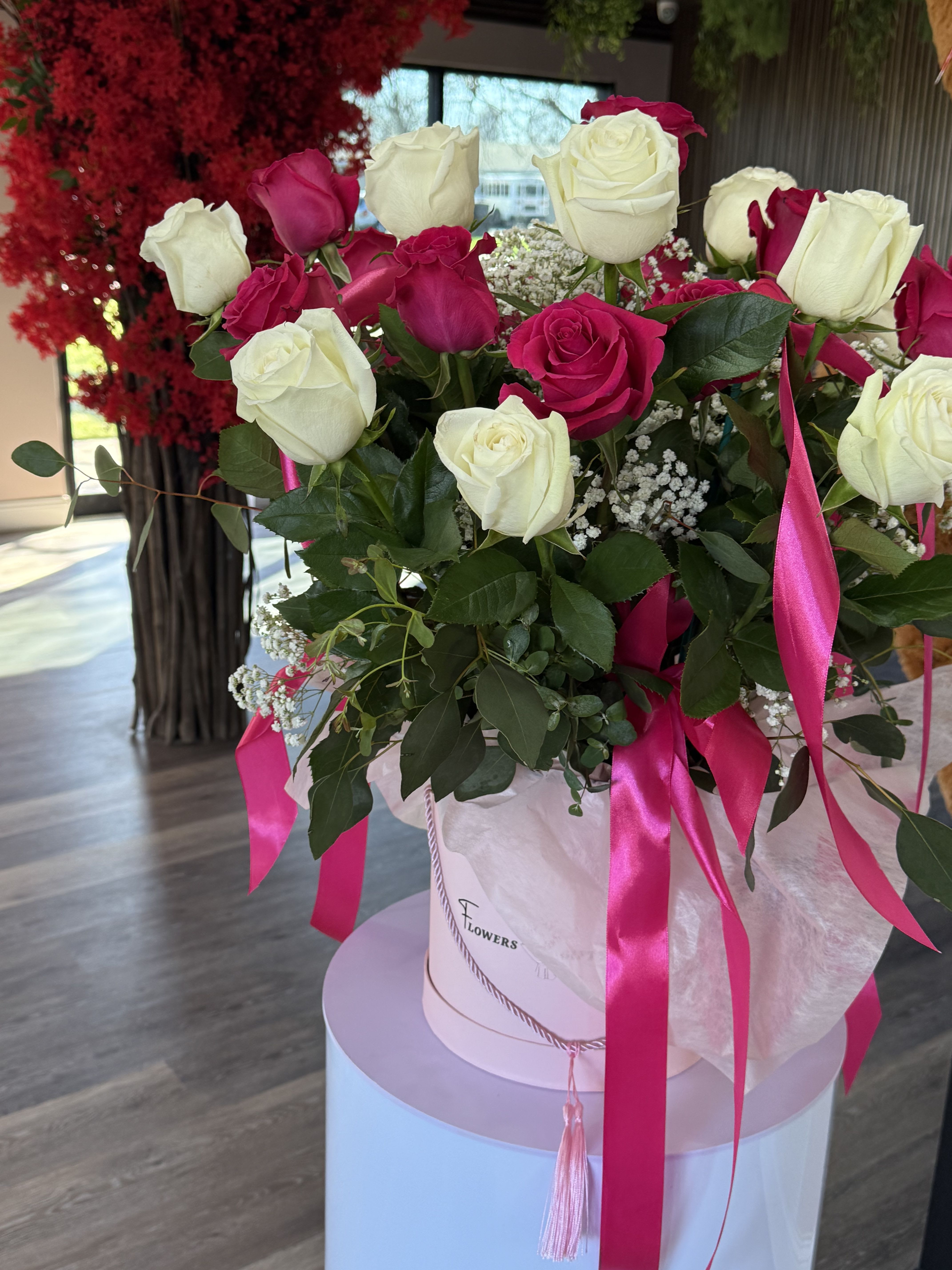 Pretty in Pink - A lush dome of white and hot-pink roses makes an immediate impression-this romantic hatbox bouquet is perfect as a birthday bouquet or love-themed gift that says "you're cherished." The crisp white roses against vivid pink blooms and airy white filler flowers create a lively, celebratory contrast that will make the recipient feel adored and truly seen.  Arranged densely in a round white or pale-pink hatbox, the bouquet is finished with long pink satin ribbons, decorative cord, and soft tissue for a polished, gift-ready look. Some versions tuck a cuddly teddy bear behind the blooms for a playful, affectionate touch. The compact, rounded shape keeps the display neat on a table or mantle and arrives ready to present-no extra wrapping needed.