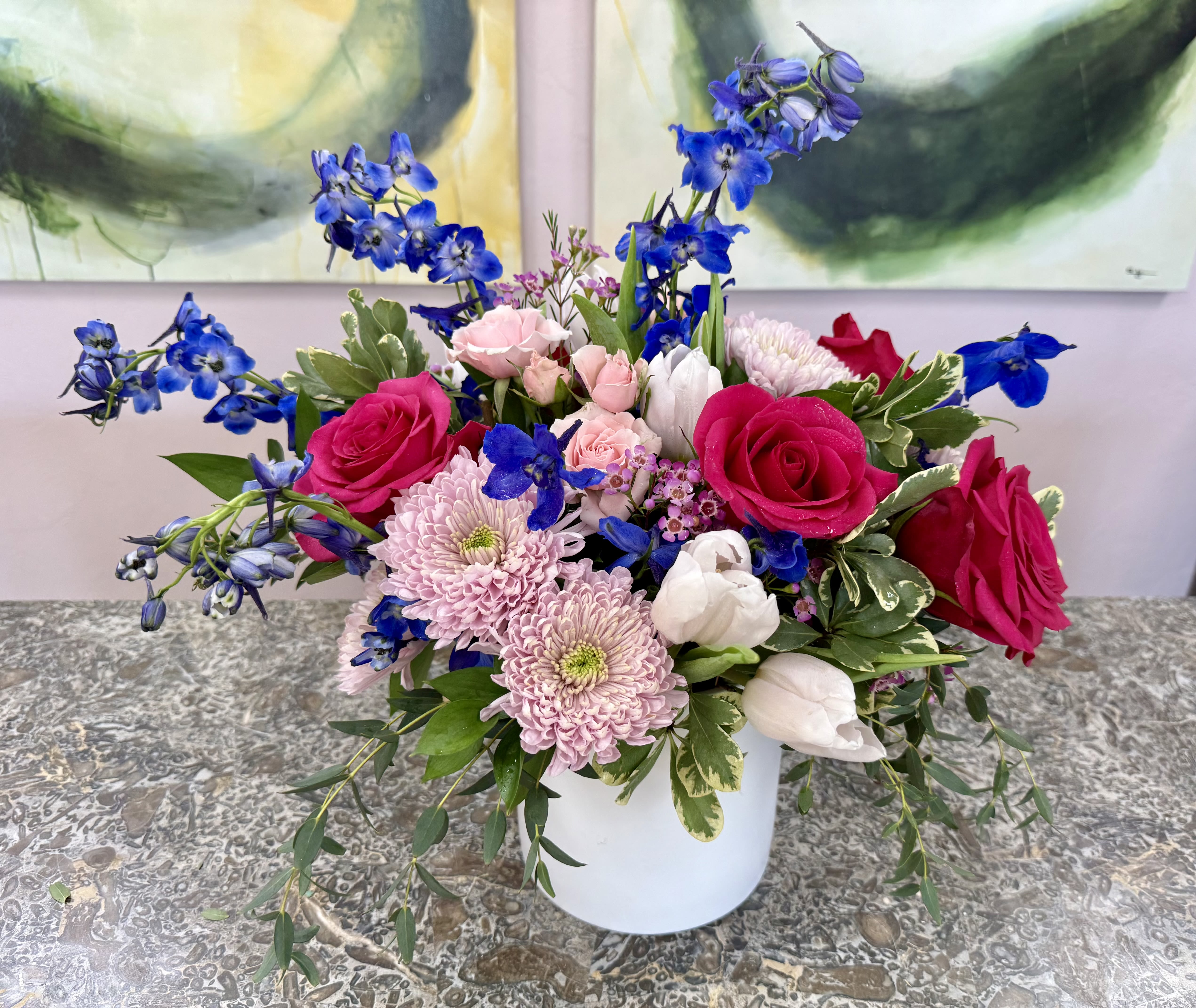 Blush and Blue - Vivid pink and soft blush roses set against cool, striking blue blooms give this flower arrangement a lively, romantic look-perfect as a birthday bouquet, anniversary roses, or an every day gift that feels effortless and personal. The blend of pinks, blues, and crisp white creates an eye-catching contrast that will make the person you're thinking of feel seen and celebrated.  This full, garden-style arrangement pairs pale pink chrysanthemums and white tulips with mixed roses and blue accent flowers arranged loosely in a simple white ceramic vase. The composition is intentionally asymmetrical, with several stems extending outward for a natural, freshly-picked texture. Sized to sit beautifully on a table or mantel, it arrives ready to display and brings a pop of seasonal color to any room.  *Flowers are subject to substitutions of equal value.