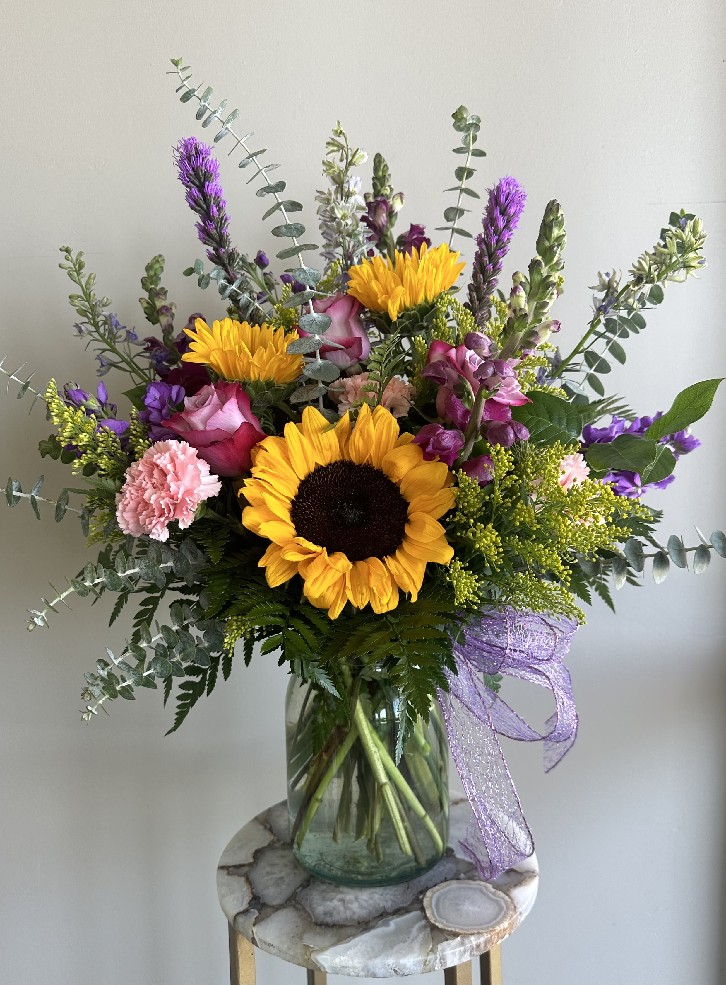 Tall mixed arrangement with sunflowers, pink roses, and purple flowers in a clear glass vase