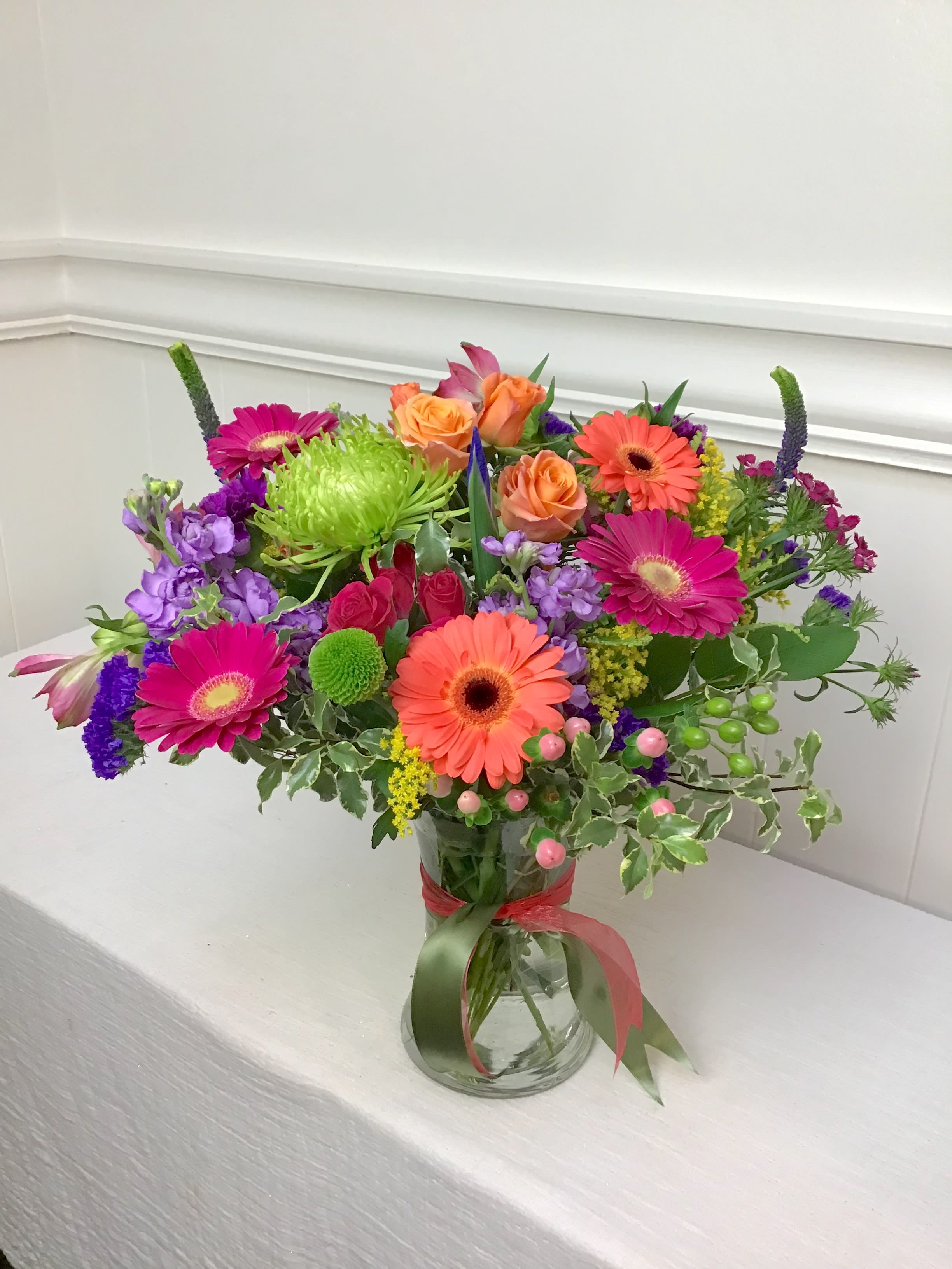 Vibrant mixed bouquet of orange roses and gerbera daisies with purple and green blooms in a clear glass vase