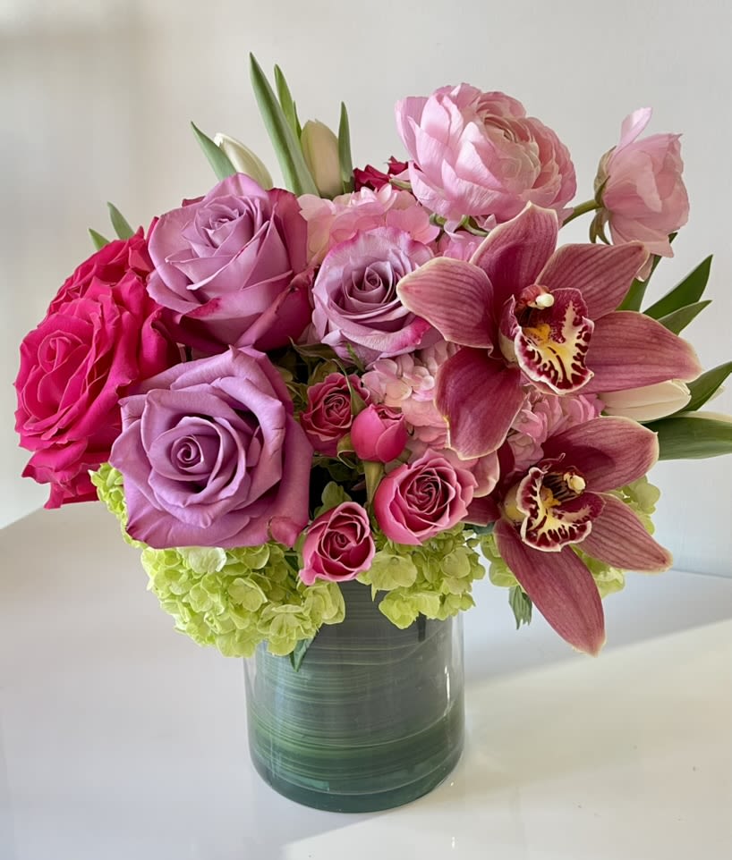 Blushing Orchid Garden - A lush, romantic arrangement overflowing with soft pinks and vibrant fuchsia tones. This elegant bouquet features velvety roses, delicate ranunculus, graceful orchids, and fresh green hydrangea, artfully arranged in a modern glass vase. Perfect for celebrating love, beauty, and life’s sweetest moments, this design brings a touch of sophistication and charm to any space.