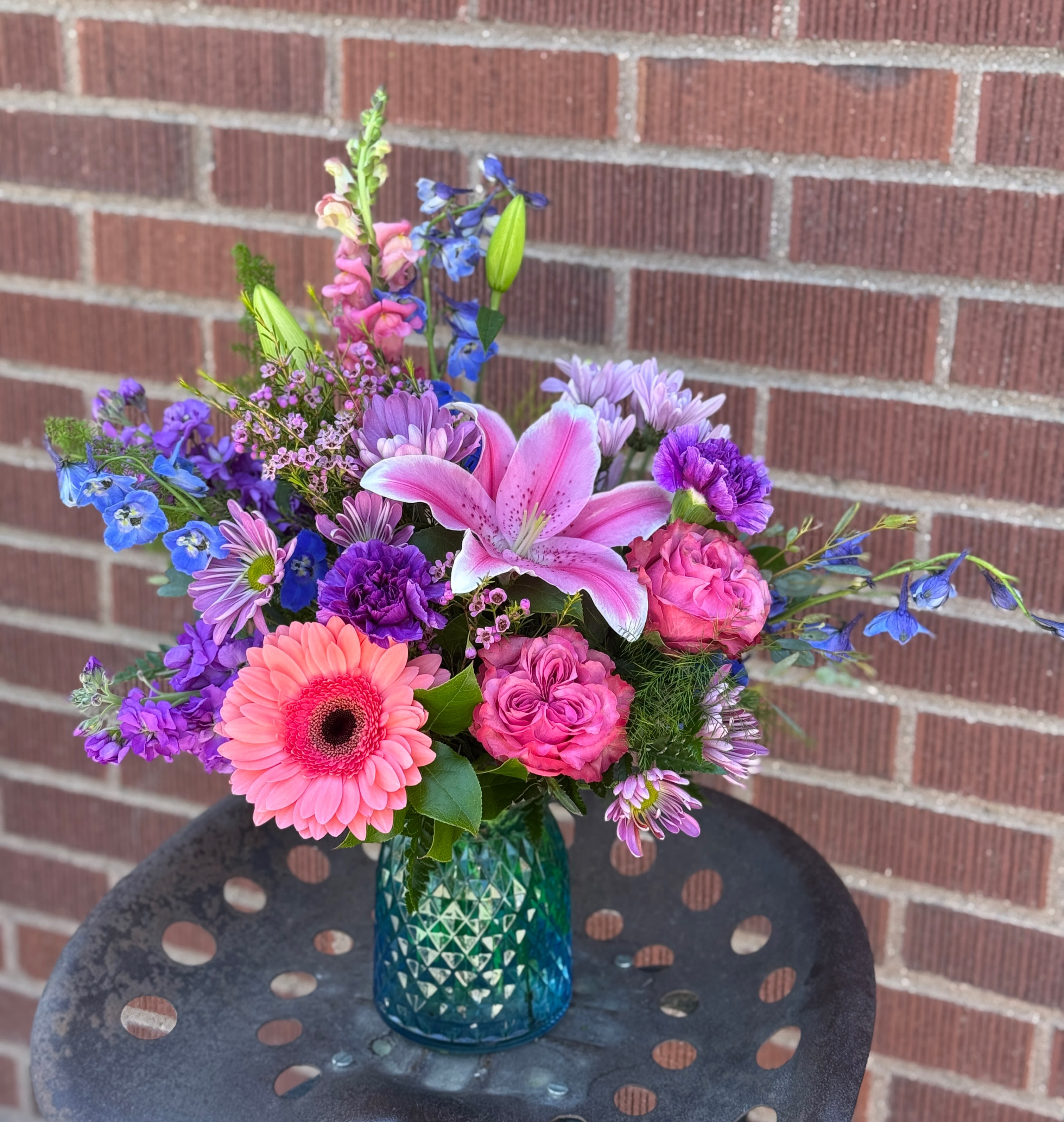 Vivid Vibrancy - A large pink lily takes center stage in this Vivid Vibrancy flower arrangement, surrounded by bright gerbera daisies, roses, snapdragons, stock and cheerful daisies in pinks, purples, blues and fresh greens. This birthday or thank-you bouquet reads like a sunlit garden-airy, asymmetrical, and slightly whimsical-making it a joyful pick for Mother's Day, spring and summer celebrations, housewarmings, or a just-because surprise that makes someone feel truly seen.  Blooming stems arrive tucked into a textured blue glass vase whose teal-to-deep-blue tones and decorative cut pattern catch the light and complement the cool color palette. The rounded body and flared rim give a modern, casual look that fits on a bedside table or metal side table-perfect for cheering a friend, saying congrats, or brightening a home. Low-maintenance and easy to display, this compact arrangement is sized for everyday gifting and ready to deliver an uplifting, colorful moment.