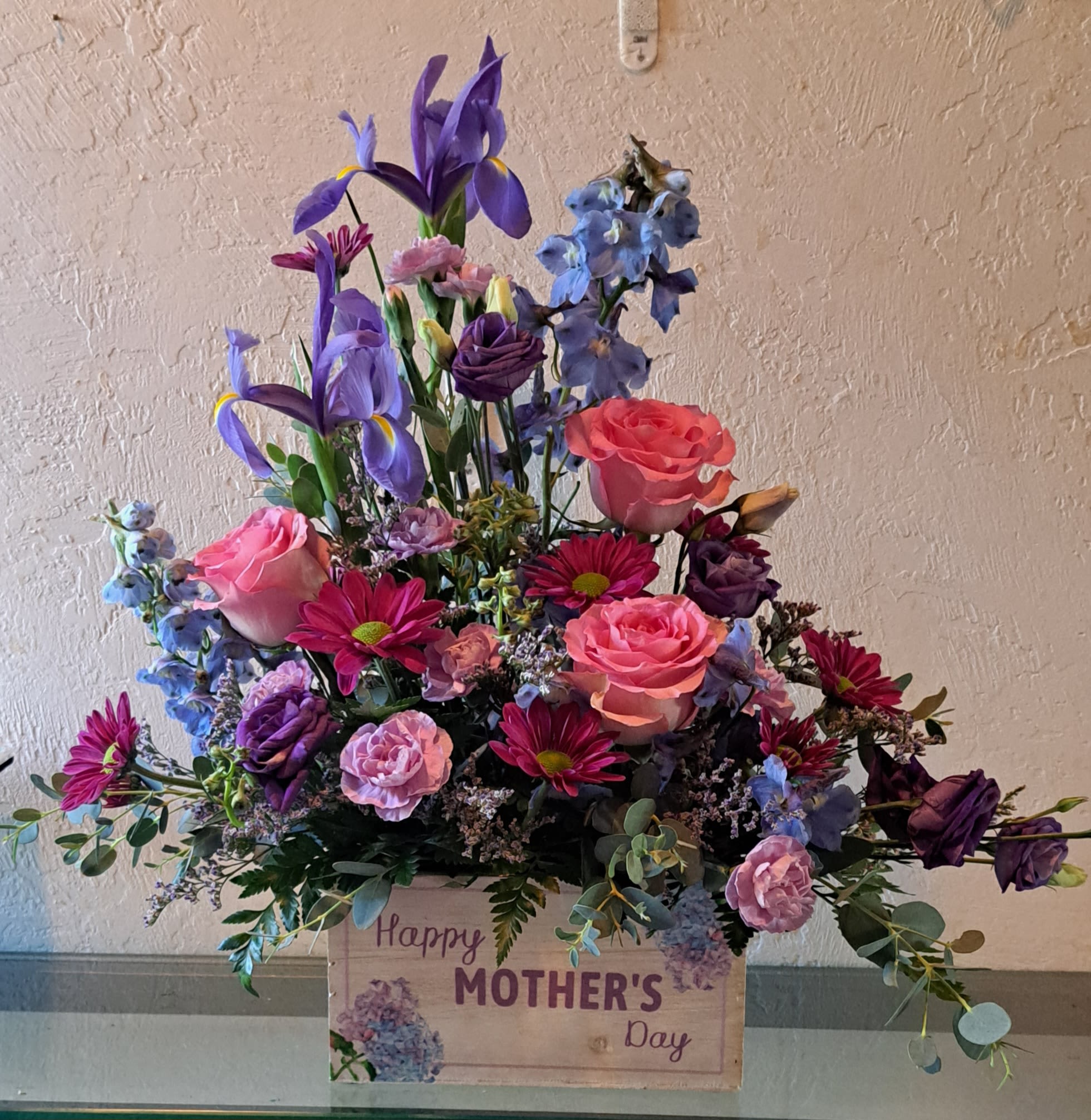 WOW Mom! - Lush layers of pink roses and purple iris create an immediate "wow" - a large, mixed flower arrangement perfect for a Mother's Day bouquet, spring surprise, or a birthday that needs to feel extra-special. Presented in a decorative gift box printed with a Mother's Day message, the arrangement reads feminine and celebratory: cool blue spikes lift the composition while warm magenta daisies and soft lavender fillers add texture and a sentimental touch.  This dense, abundant composition combines classic roses, tall iris, delphinium-like blue spikes, cheerful daisies, and delicate filler blooms for a full, garden-fresh look. The color story-pink, purple, white, and blue-balances vibrancy with softness so she'll feel both celebrated and cherished. Sized as a large, layered bouquet in a keepsake box, it arrives ready to display and makes a thoughtful "thank you," "thinking of you," or springtime gift.