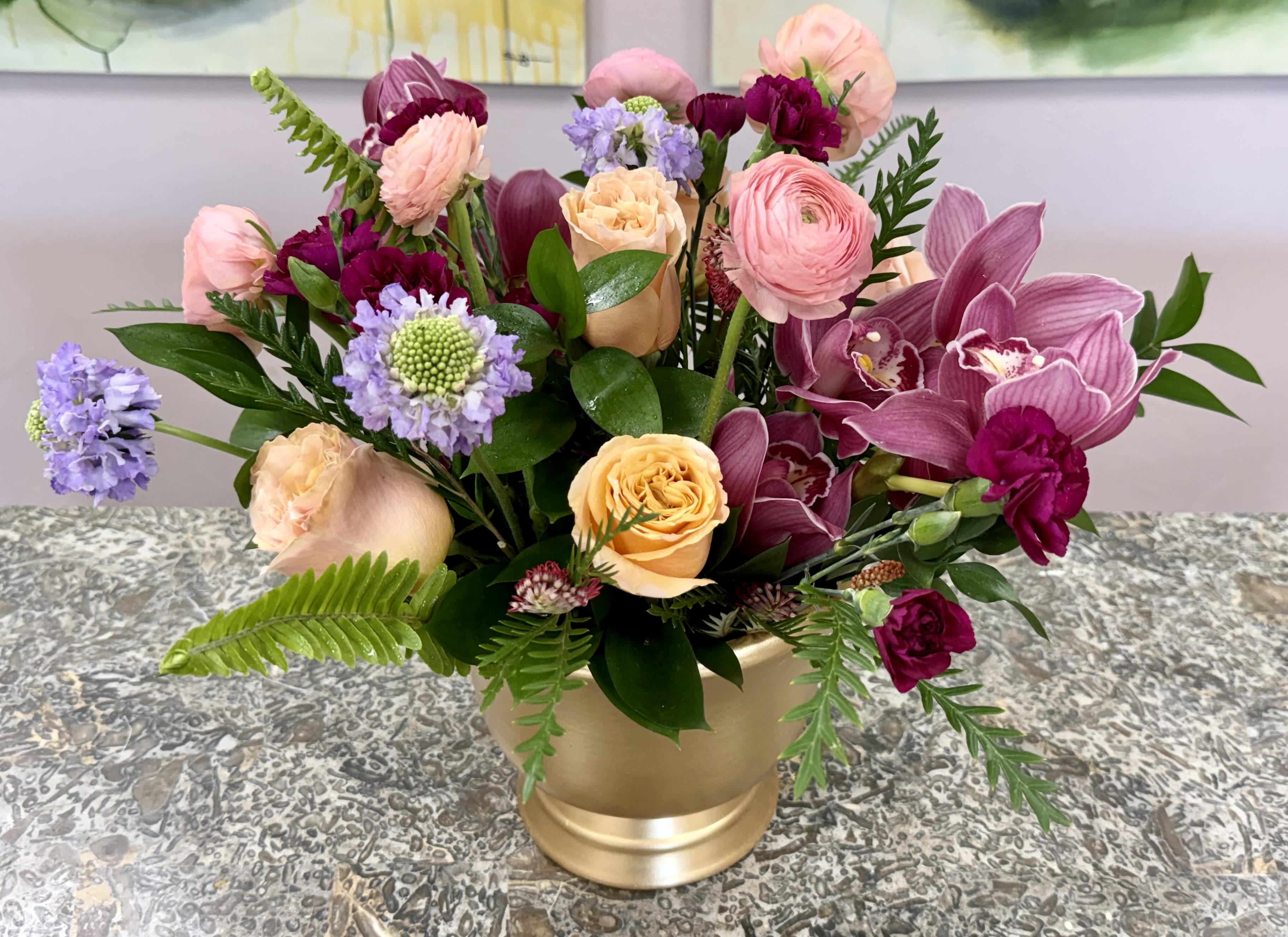 Peach and Amethyst Luxe - Blush, peach, magenta and lavender blooms mingle in a lush, rounded design that feels both romantic and celebratory-perfect as an anniversary bouquet, birthday arrangement, or a springtime surprise for someone special. The composition highlights large orchid blooms that balance softer roses and carnations, creating a mix of bold focal flowers and velvety, comforting petals that read feminine and refined.  Smaller filler flowers add delicate texture and contrast across the arrangement, while a polished gold vase lifts the palette with a touch of luxe and formality. Sized to dress a dining table or brighten an entryway, this arrangement is crafted to make the recipient feel truly seen and celebrated; choose it when you want to send a thoughtful, elegant statement that reads as both modern and timeless. *Flowers are subject to substitutions of equal value.
