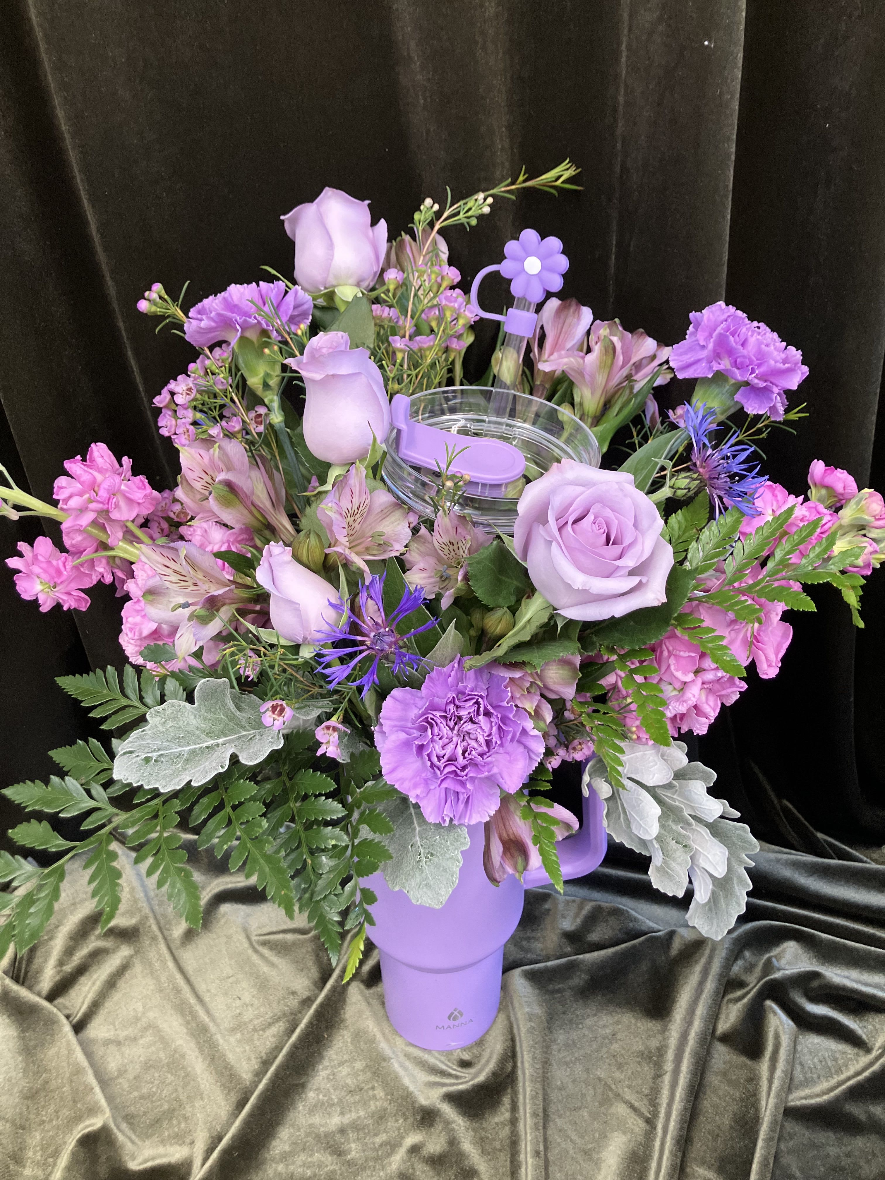 Lavender Tumbler  - Lavender roses and soft pink carnations come together in this Lavender Tumbler to create a cheerful, romantic gift - an ideal birthday bouquet, Mother's Day surprise, or just-because pick-me-up. The rounded, abundant composition reads feminine and playful; the recipient will feel noticed and cherished when they lift the insulated purple tumbler to reveal the blooms inside.  Petals include lavender roses, pale pink carnations, delicate alstroemeria, and small blue accent blooms set against silvery foliage and fern-like greens for a soft, textured look. Flowers arrive arranged in a purple plastic insulated tumbler with a clear lid and a fun plastic straw topper, making the presentation both giftable and reusable as a keepsake. Lightweight and compact, it's ready to display on a desk, bedside table, or kitchen counter - fresh water and a cool spot away from direct sun will help the stems last.