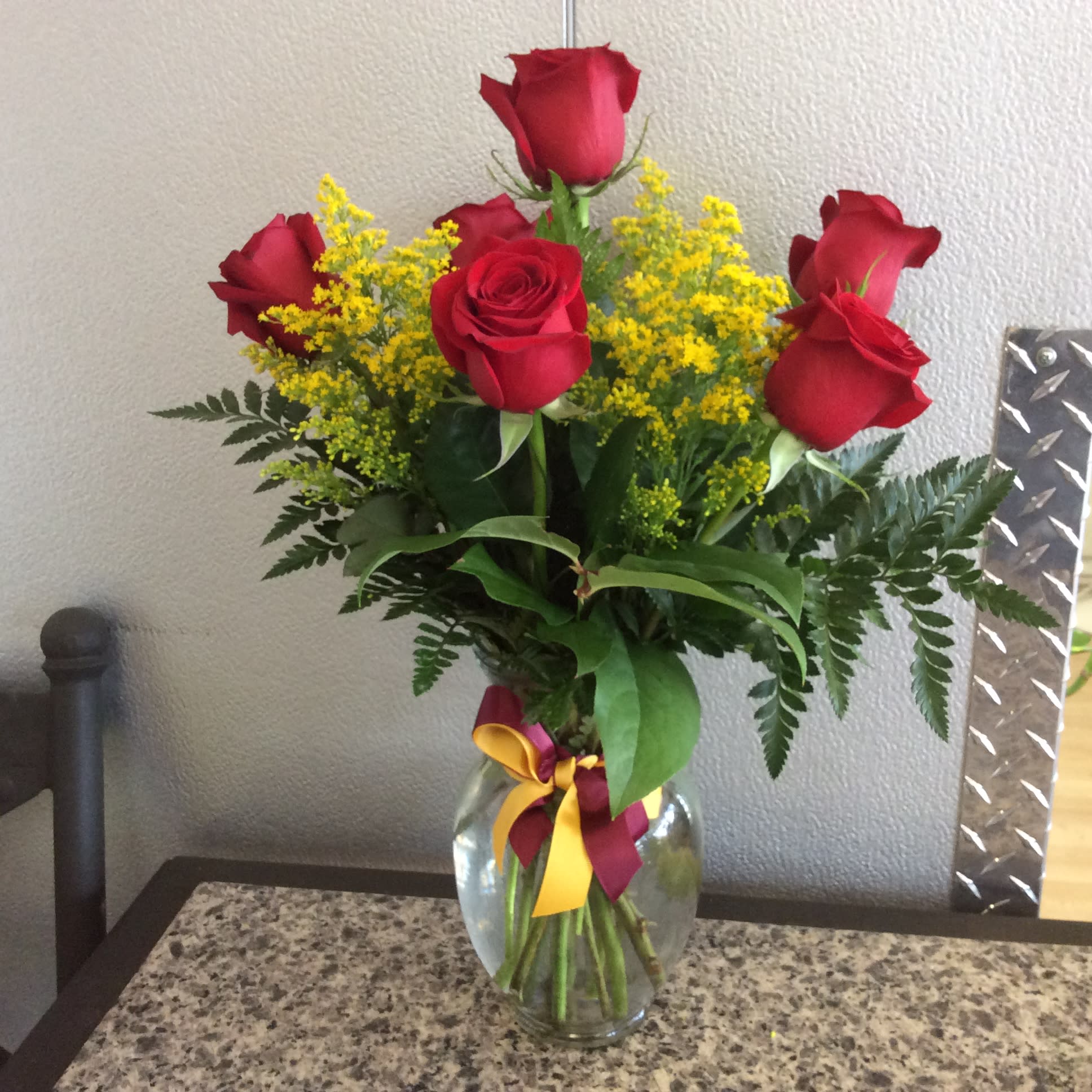 Arrangement of red roses with yellow filler flowers in a clear glass vase with a burgundy and yellow bow
