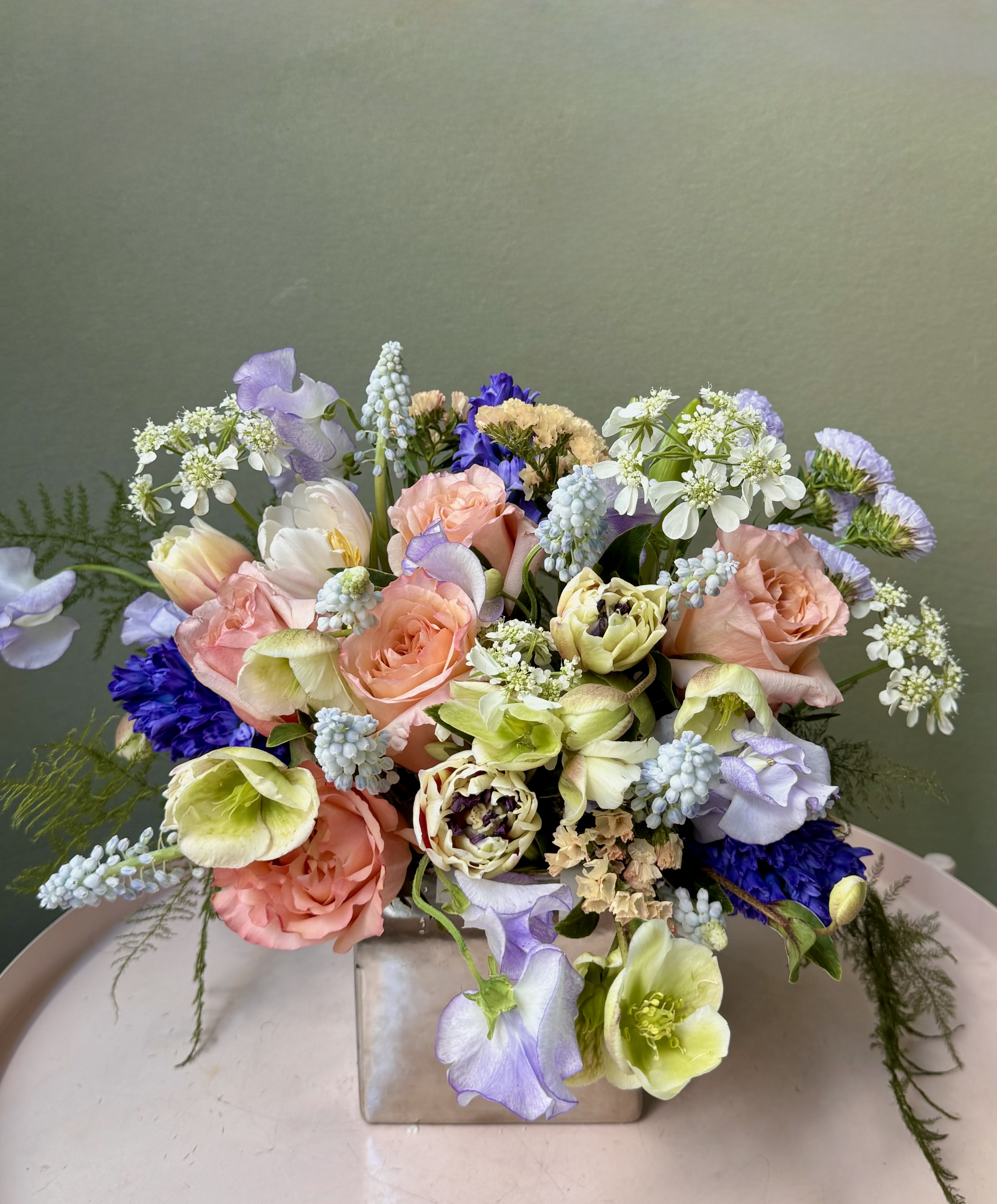 Lauren  - Soft peach and pink roses mingle with lavender and white tulips in a pastel bouquet designed to feel like a sunlit garden-perfect as a birthday bouquet, spring gift, or delicate wedding table arrangement. The Lauren flower arrangement sends a gentle, romantic message the person you're thinking of will notice right away: soft colors, airy texture, and a loose, rounded shape that reads intimate and effortless.  This composition pairs pale blue grape hyacinth and small white daisy-like blooms with mixed greenery for light contrast and movement; a few trailing stems extend beyond the main mass for a natural, garden-picked look. Arranged in a clean square ceramic vase, Lauren measures as a compact yet full centerpiece that's easy to display on a mantel, bedside table, or reception table-an elegant way to make someone feel truly seen.