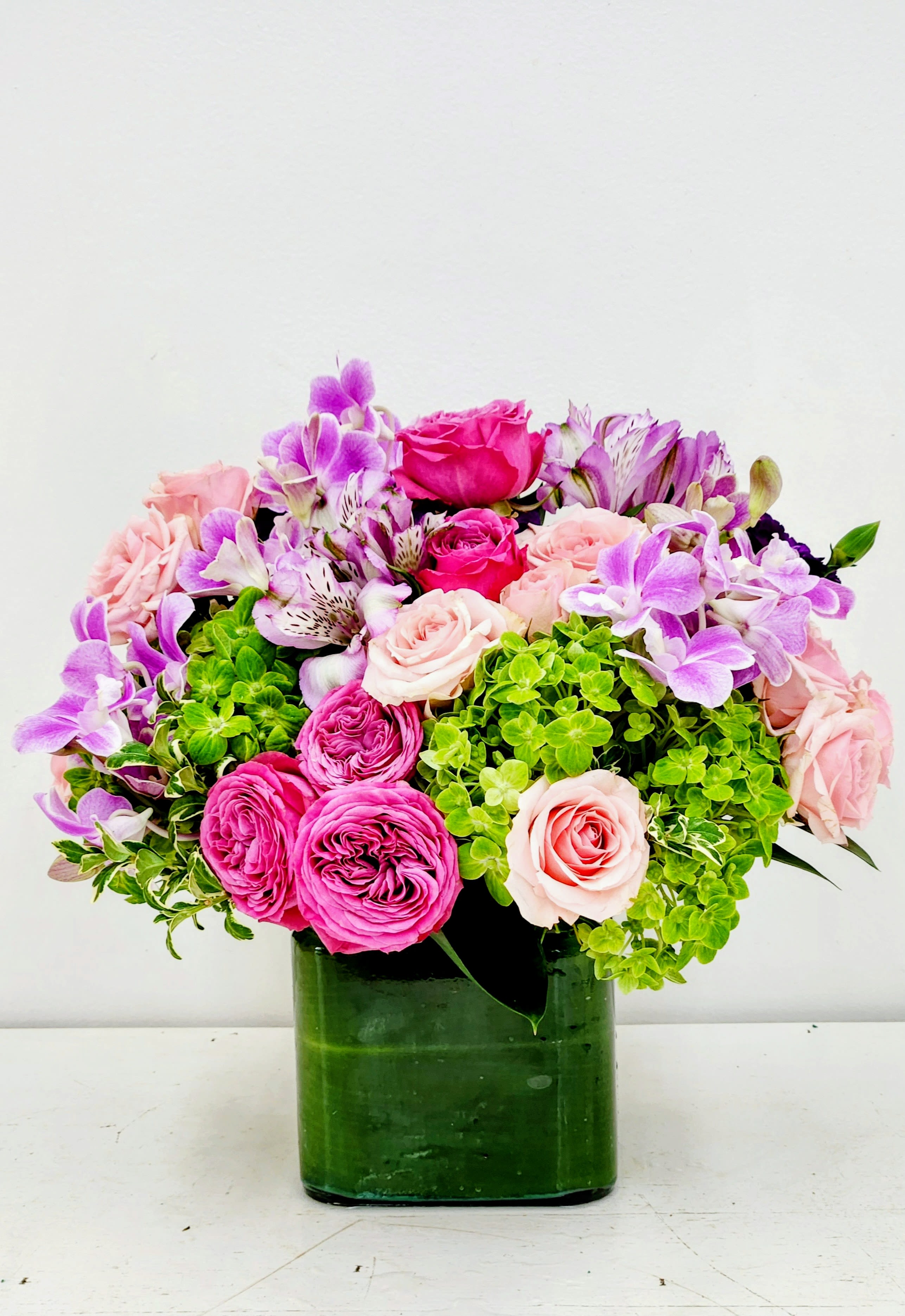 Beautiful and Blooming - Hot pink and pale blush roses mingle with lavender alstroemeria and pops of purple in a compact, rounded flower arrangement-perfect as a Mother's Day bouquet, a thoughtful Thank You gift, or a warm Thinking of You surprise. The bright green hydrangea clusters give the design a vivid contrast that reads joyful and romantic, so the person you're sending it to will feel celebrated and seen the moment the vase arrives.  This lush arrangement is built low and full, layering roses, alstroemeria, and hydrangeas for a dense, garden-fresh texture with a polished contemporary silhouette. Finished in a dark green glass vase that anchors the saturated palette, it reads elegant on a dining table or brightens an office desk. Ideal for birthdays, anniversaries, or congrats moments, it arrives ready to display; refresh with fresh water and trim stems every few days to keep blooms looking their best.