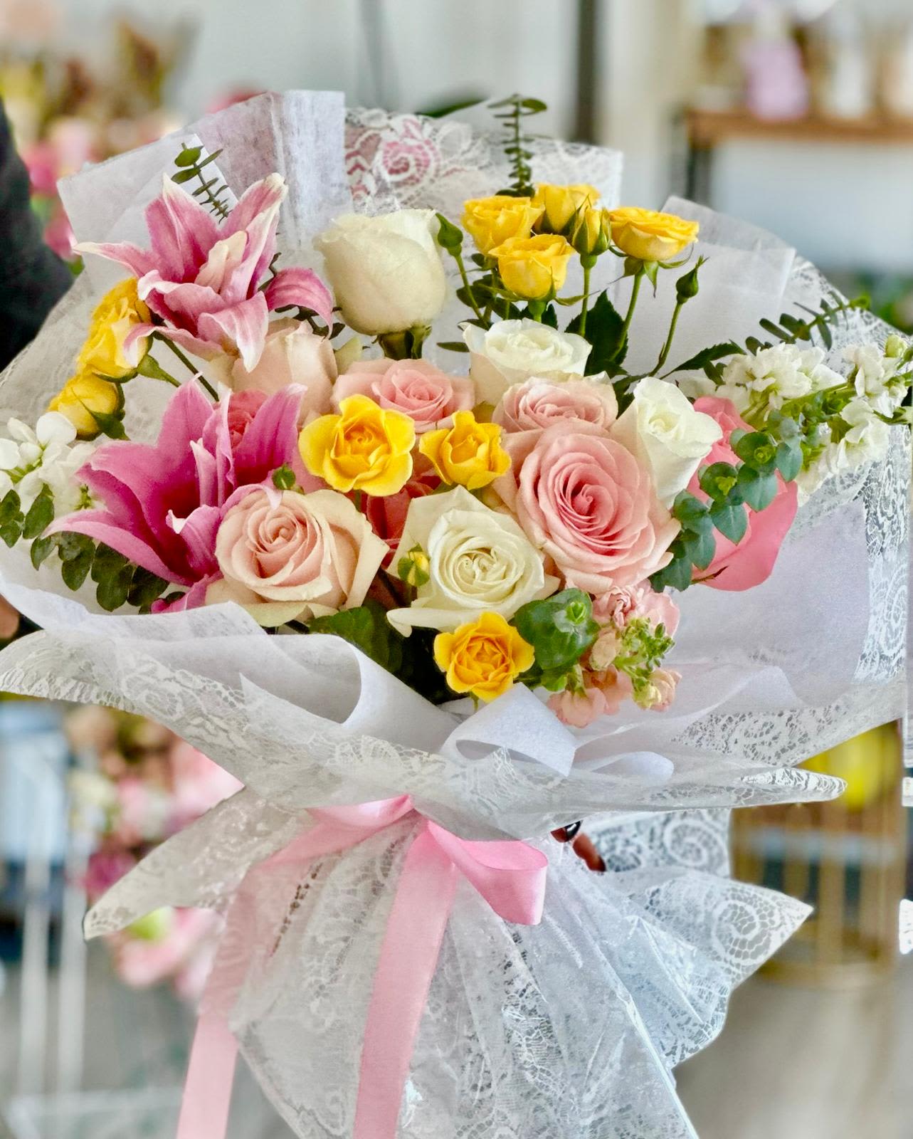 Lace Bloom  - A round bouquet with pink, white, and yellow flowers, wrapped in lace paper and clear wrapping.   Flower types may vary depending on the season.