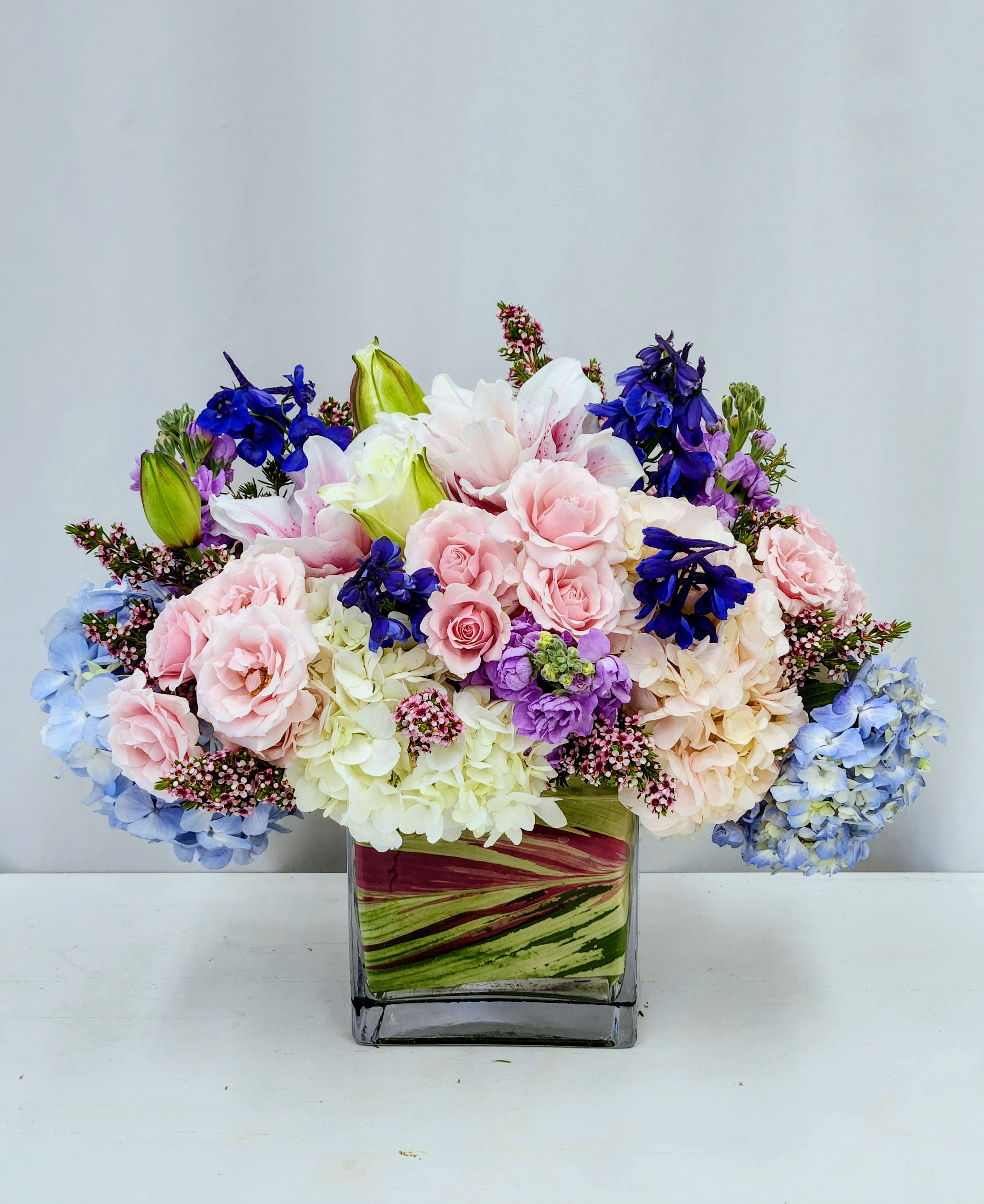 Calming Beuaty - Soft pink roses paired with pale and blue hydrangeas create an airy, romantic centerpiece that feels both celebratory and quietly soothing - perfect as a birthday bouquet, anniversary arrangement, or spring gift to show someone you're thinking of them. The mix of white lilies and vivid blue and purple accent blooms gives the Calming Beauty arrangement a layered, garden-fresh look that will make the recipient feel cherished and seen.  