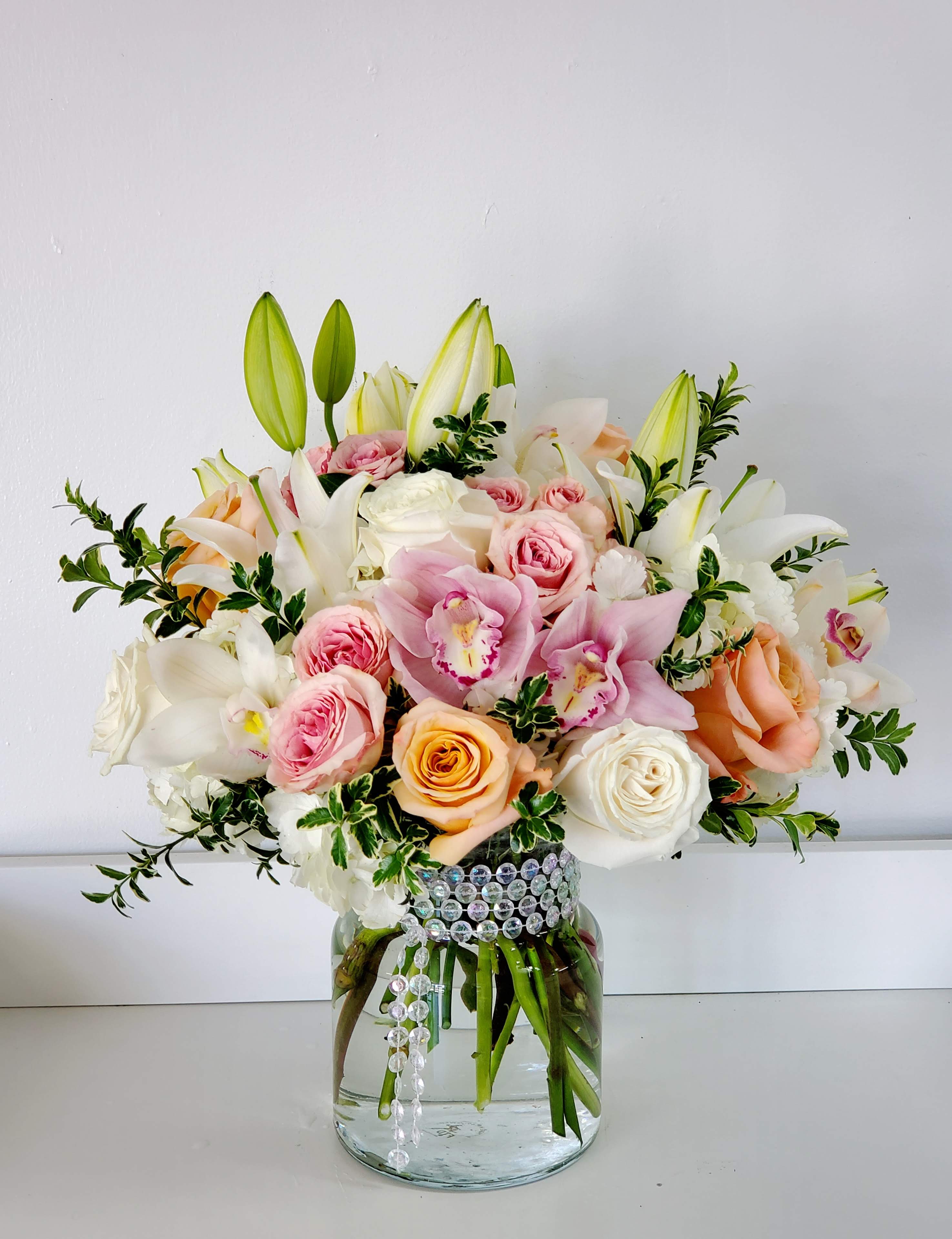 Dainty - Soft pastel roses, white lilies, and delicate pink orchids come together in a rounded, full bouquet that reads like a quiet celebration-perfect as a birthday bouquet, anniversary arrangement, or Mother's Day gift. The mix of pink, white, peach, and fresh green tones creates a romantic, elegant mood that will make the person you're thinking of feel cherished and seen.  