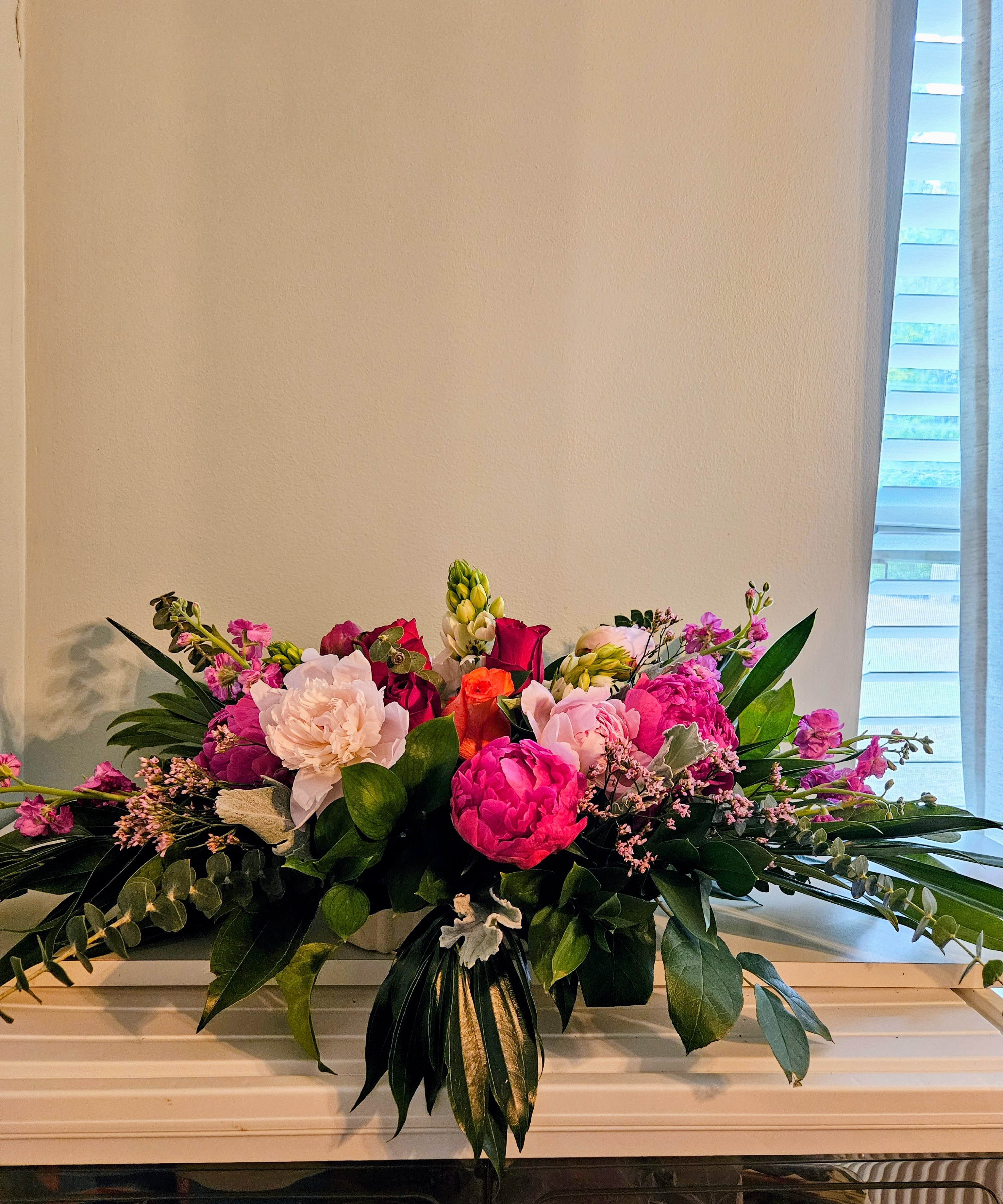 " Fortune Waltz" - A lush, low centerpiece that reads like a celebration in bloom, perfect as a birthday bouquet, anniversary arrangement, or Mother's Day gift. This flower arrangement places large pink peonies and pale blush blooms front and center, with vivid red roses weaving through soft pink carnations and stock to create a romantic, feminine statement the recipient will notice the moment they enter the room.  Arranged in a rectangular container, the design spreads horizontally for a full, abundant silhouette-layered greenery and long leaves extend outward to give the piece movement and texture. White accents lift the palette and deepen the contrast against rich green foliage, while smaller pink filler flowers add delicate detail around the larger blossoms. The overall effect is bright, elegant, and intentionally garden-fresh.  Ideal for the person you want to make feel celebrated, seen, or loved-this centerpiece works well on dining tables, mantelpieces, or as a thoughtful everyday surprise. Care is straightforward: keep water fresh, trim stems every few days, and display out of direct heat to prolong the life of the blooms.