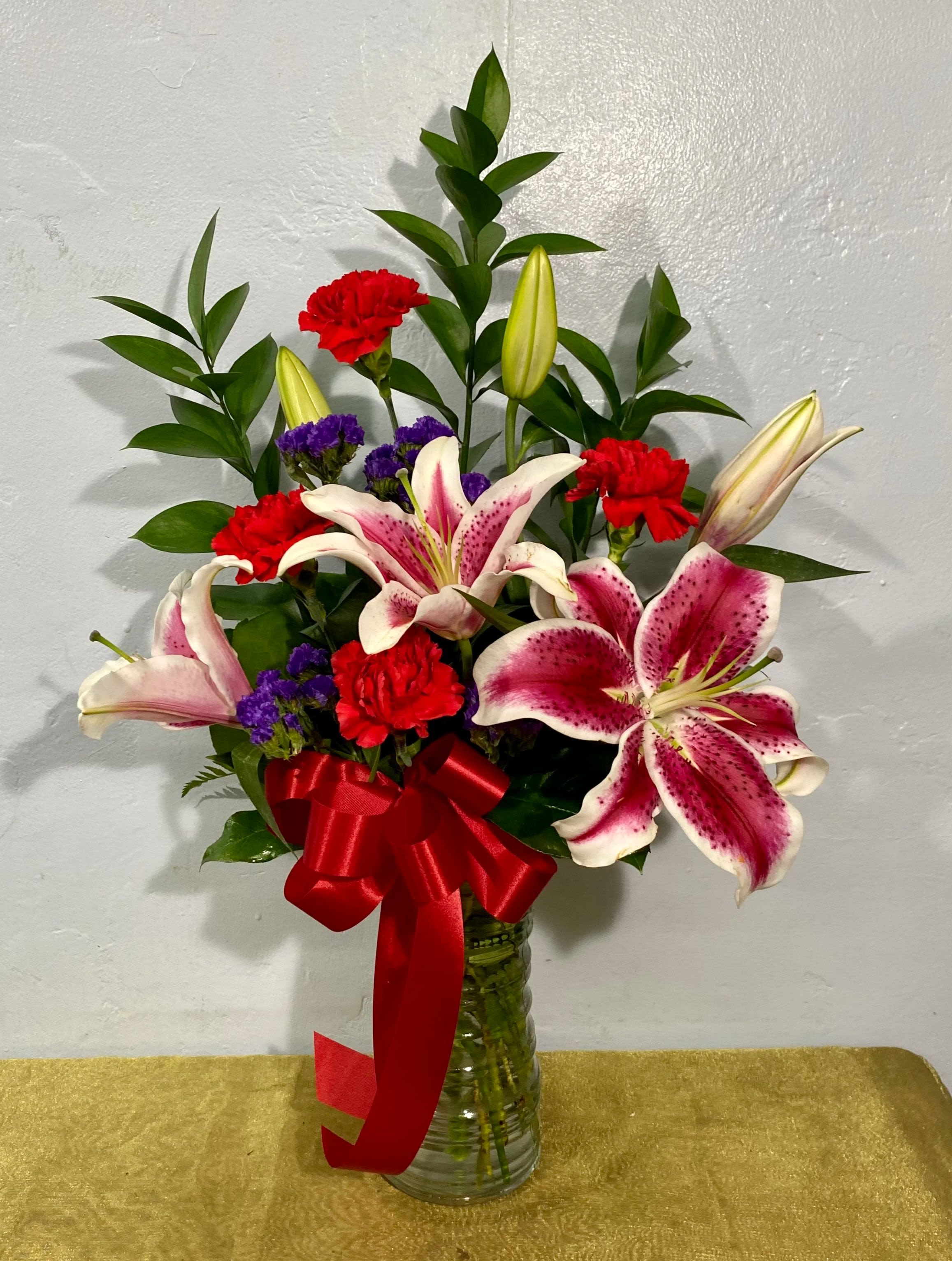 Lovely Lilies - These amazing lilies are sure to delight!  Beautiful pink lilies, red carnations, filler flowers and foliage hand arranged in a clear cylinder vase.  Approximately 25" tall and 18" wide.