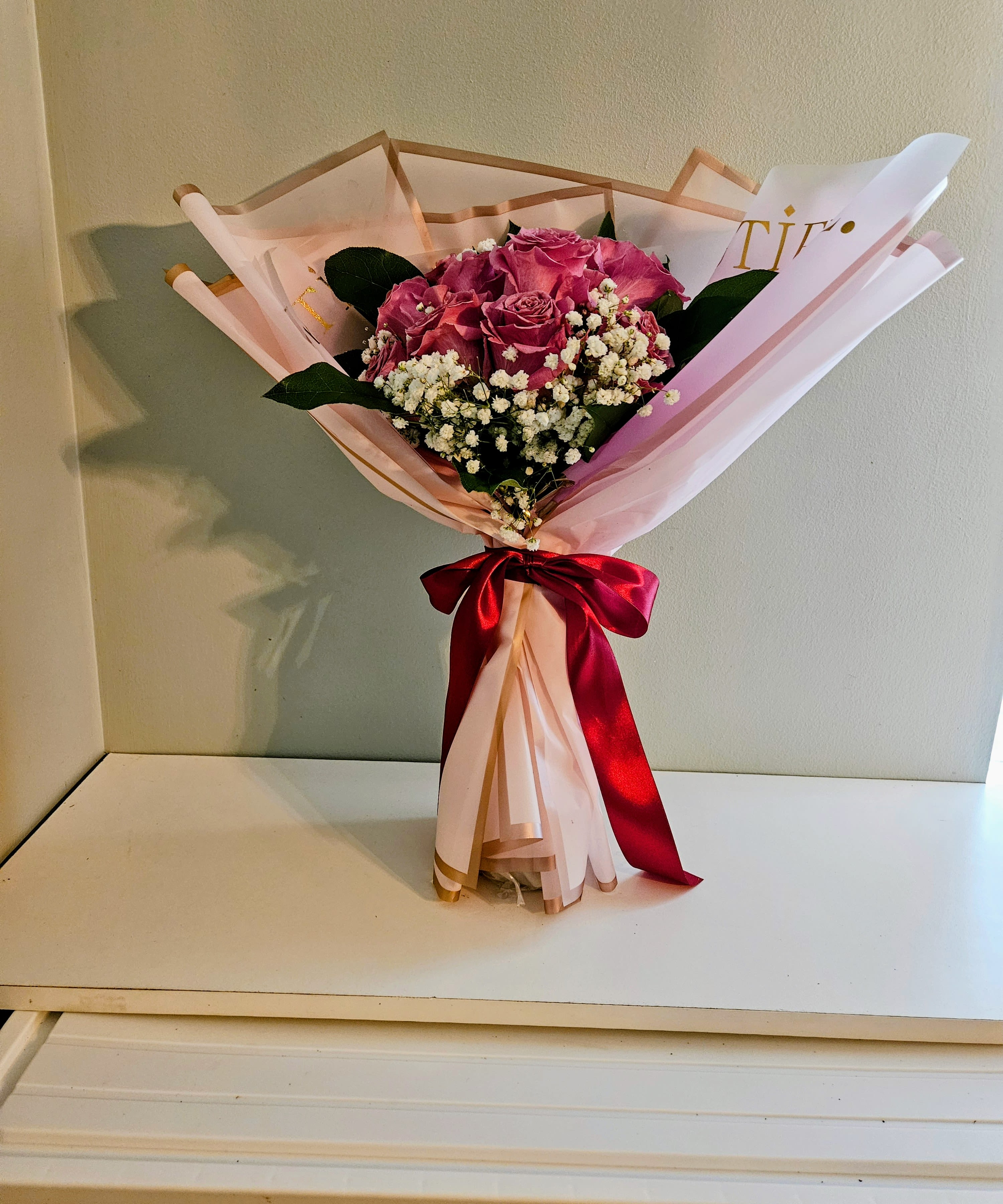 " Tower of Purple" - A cluster of soft pink roses takes center stage in this flower arrangement, making the Tower of Purple an elegant birthday bouquet or anniversary roses choice that quietly says "I love you." The close-set pink blooms, lifted by small white filler flowers and deep green leaves, create a romantic, feminine impression that suits celebrations, thank-you moments, or a thoughtful just-because gift for someone you want to make feel seen.  Wrapped in pale pink decorative paper edged in gold and finished with a deep red satin ribbon, the presentation feels polished and gift-ready. The mix of pink roses, white accents, and rich foliage gives a lush, tactile look-soft petals against glossy leaves-that reads classic and slightly formal. Sized for tabletop display, it arrives ready to place in a vase; trim stems and refresh the water every few days to keep the roses at their best.