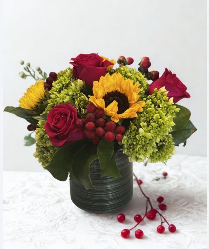 Sunset Pedals - This vibrant, textured arrangement is the perfect way to celebrate a special occasion. Featuring red roses, and cheerful sunflowers balanced with green hydrangeas and pops of hypericum berries. tucked in a glass vase