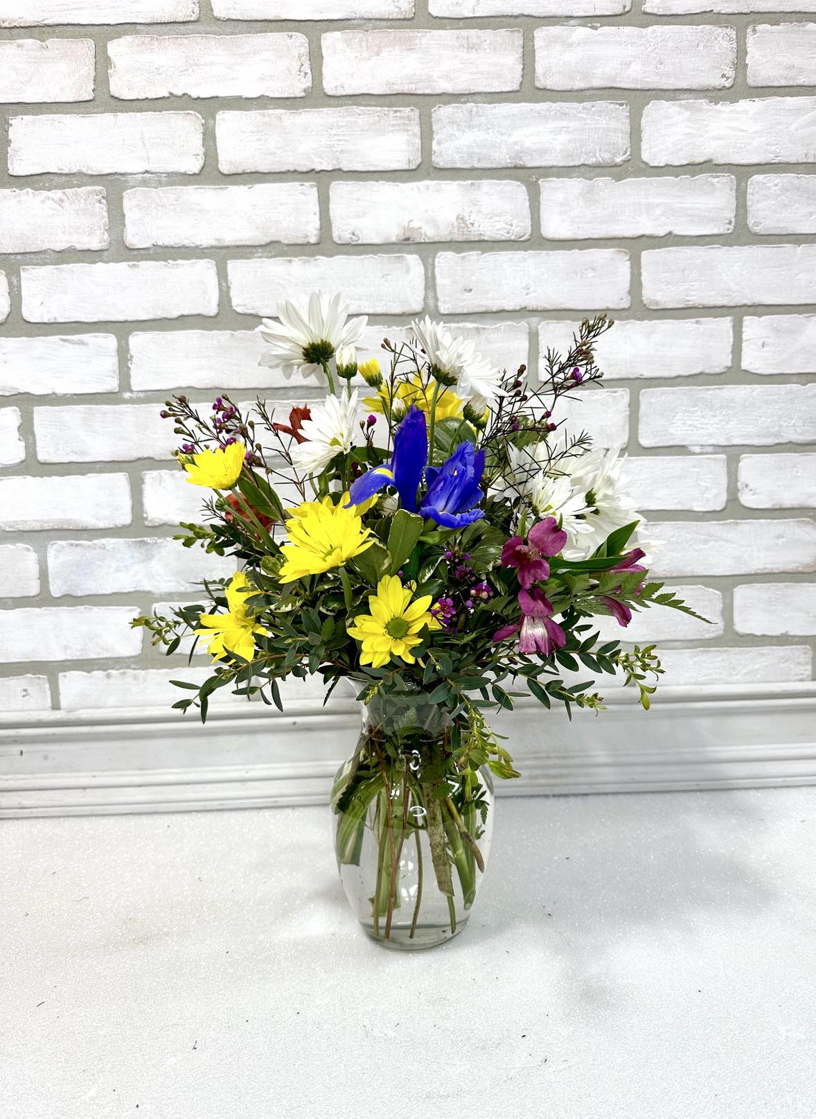 Mixed bouquet of yellow, white, blue, and purple flowers in a clear glass vase