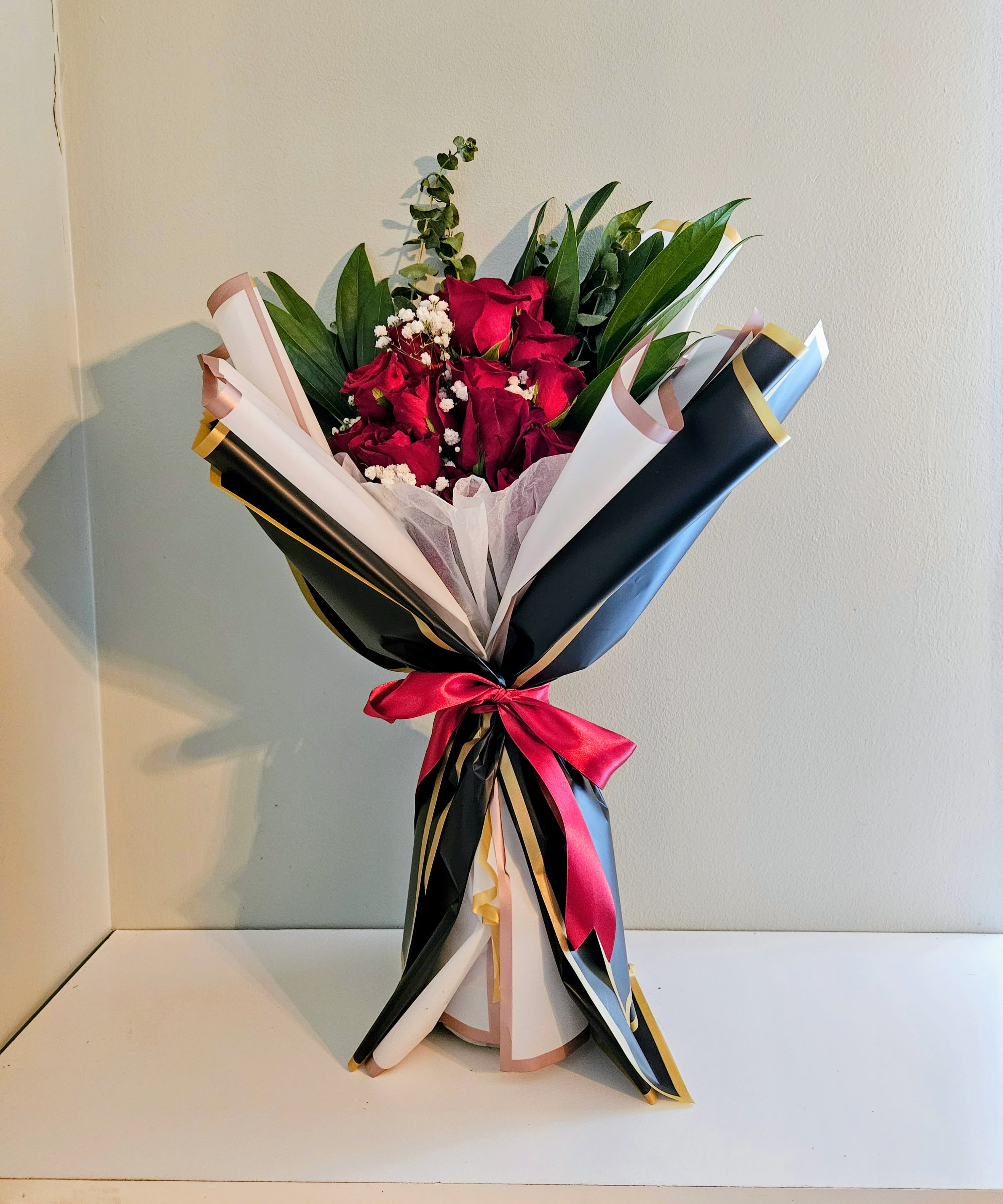 "Towering Of Romance" - A dense cluster of deep red roses commands attention in this romantic flower arrangement - the perfect anniversary roses or love-and-romance bouquet to make someone feel truly cherished. The dramatic contrast of red blooms against black, white, and blush layered wrapping gives it a polished, gift-ready look that also suits birthdays or a heartfelt thank-you.  Small white filler flowers and upright greenery add texture and a fresh, sculpted silhouette, while gold-edged paper and a bright red satin ribbon heighten the luxe presentation. Finished with decorative wrapping paper, this bouquet reads formal and elegant when held or displayed on a table.  Designed to arrive as a memorable, ready-to-give gift, Towering Of Romance is ideal for the person you want to sweep off their feet. Keep blooms in fresh water and away from direct heat; for best enjoyment, recut stems and change water every few days to extend vase life.