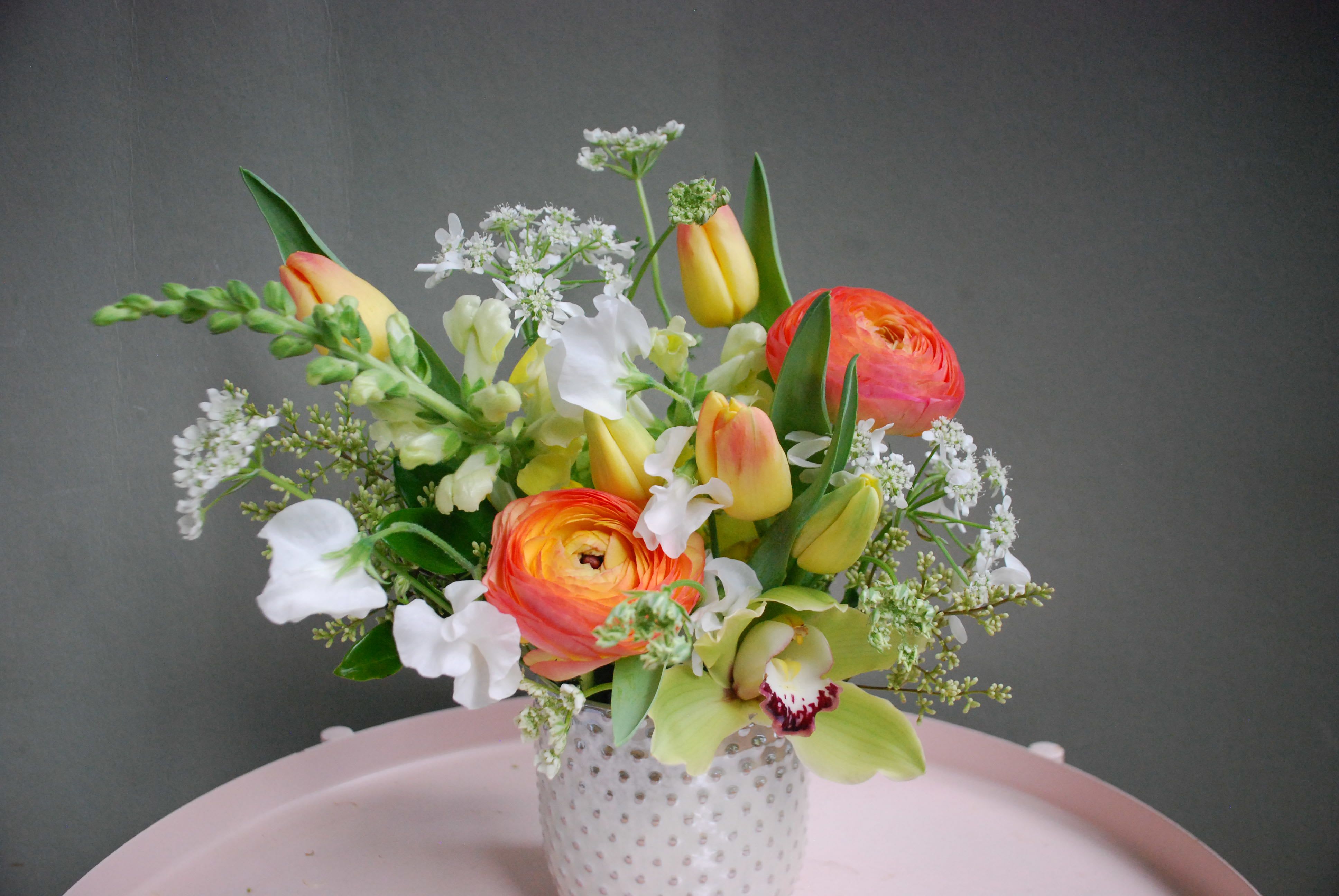 Josephine  - Bold, sculptural and joyfully colorful, this floral arrangement-perfect as a birthday bouquet or special-occasion centerpiece-pairs dramatic tropical blooms with airy stems to make the recipient feel truly celebrated. The design reads like modern floral art: tall, vivid red tropical flowers and round purple allium-like heads create striking vertical interest, while cascading purple orchid sprays and pops of mixed-color blooms bring movement and a celebratory energy suited to anniversaries, milestone events, or an upscale gift.  On a closer look, Josephine blends seasonal favorites-tulips, ranunculus, orchids and delicate filler flowers-in palettes that range from soft pinks, yellows and oranges to bold reds, purples and fresh greens. One presentation sits in a white textured ceramic vase that adds clean, contemporary contrast to the loose, garden-style composition; other arrangements showcase more sculptural groupings on a dark tabletop for an event-ready statement. The mix of open tulips and layered ranunculus gives a lush, tactile feel while small white accents brighten the overall look.  Choose Josephine when you want someone to feel seen-whether it's a springtime birthday, a cheerful everyday surprise, or a dramatic display for a special event. Sized to suit tabletops or mantels, the textured ceramic vase option offers a modern keepsake; to keep blooms looking their best, change the water every couple of days and recut stems at an angle when arranging.