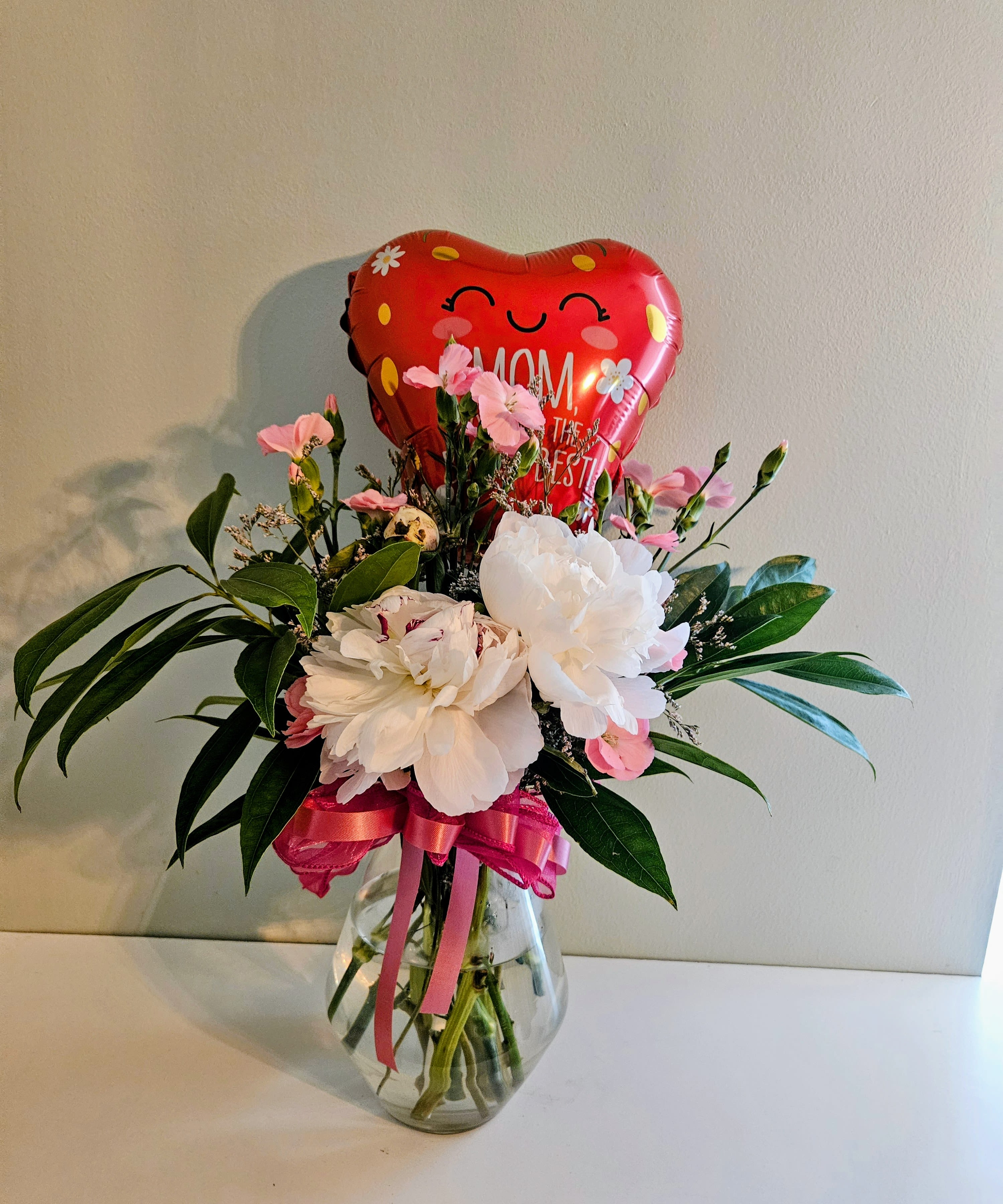 Prosperity Of Living - Soft pink and white carnations paired with glossy green foliage create a gentle, joyful statement perfect as a Mother's Day bouquet or a heartfelt appreciation gift. The arrangement reads as playful and feminine-the red heart-shaped foil balloon with a smiling face and Mother's Day message lifts the design literally and emotionally, making the recipient feel celebrated and seen.  Carefully arranged in a clear glass vase and finished with a pink ribbon, this flower arrangement blends white and pink carnations with small pink accent blooms for a layered, textured look. The glass vase keeps the focus on the blooms and foliage while the balloon adds height and a cheerful pop of red. Size and exact flower counts vary by selection; vase is included.  This is the kind of gift that brightens a kitchen table, office desk, or bedside stand and makes Mom or a special woman in your life feel genuinely loved. Easy to care for-keep fresh water in the vase, trim stems if needed, and place away from direct heat-to extend the life of the carnations and foliage.