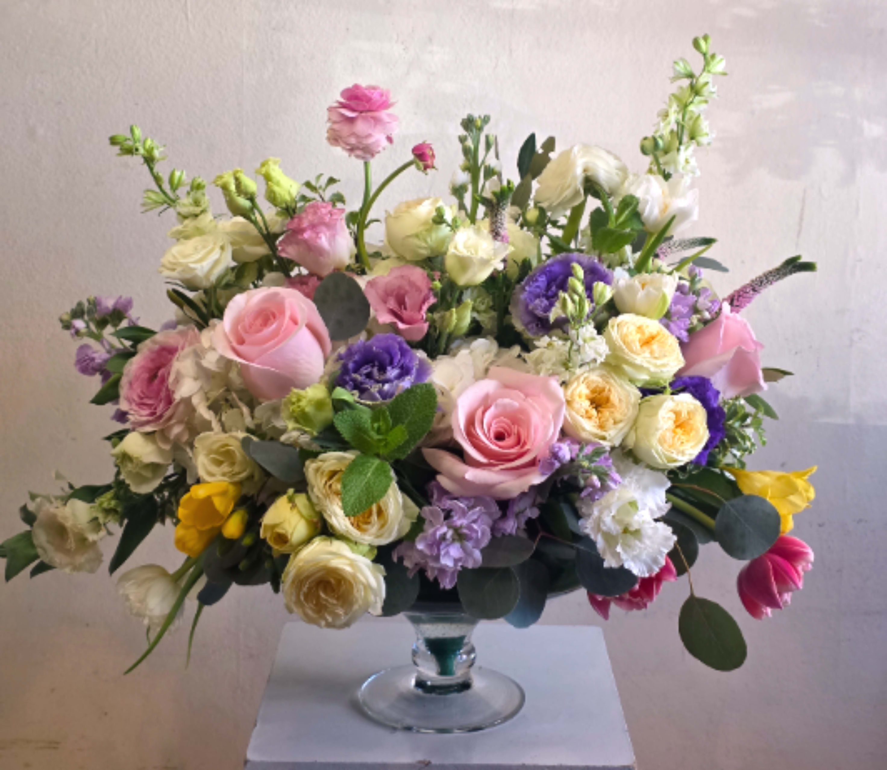 Spring Garden - A lush, garden-fresh composition of mixed bouquets in soft pastels-this Spring Garden flower arrangement brings together roses, spray roses, lisianthus, hydrangea, ranunculus, tulips, freesia and stock in a rounded, airy display perfect for a spring birthday bouquet, Mother's Day gift, anniversary roses, or a wedding centerpiece. The mix of pink, white, lavender, purple and sunny yellow creates a gentle, romantic feeling that makes the person you're thinking of feel truly celebrated-whether you're sending love and romance, a thank-you, or a get-well wish.  Arranged in a clear glass pedestal vase or pedestal bowl, the design is lush and textured: large pink roses and creamy yellow spray roses anchor clusters of pale hydrangea and stock, while lavender lisianthus and deeper violet accents add depth. Bright yellow and pink tulips and delicate freesia bring lively pops of color and a garden-like movement; dimensions and bloom count vary by size, and upgraded orders receive additional blooms in a larger vessel for extra impact.  Ideal for Spring and Easter gatherings but lovely any time of year, Spring Garden also suits thinking-of-you moments, new baby celebrations, housewarmings, sympathy remembrances, or a just-because surprise. Care: keep stems in fresh water, recut stems and remove wilted foliage as needed to extend vase life; select your preferred size at checkout to match the occasion.