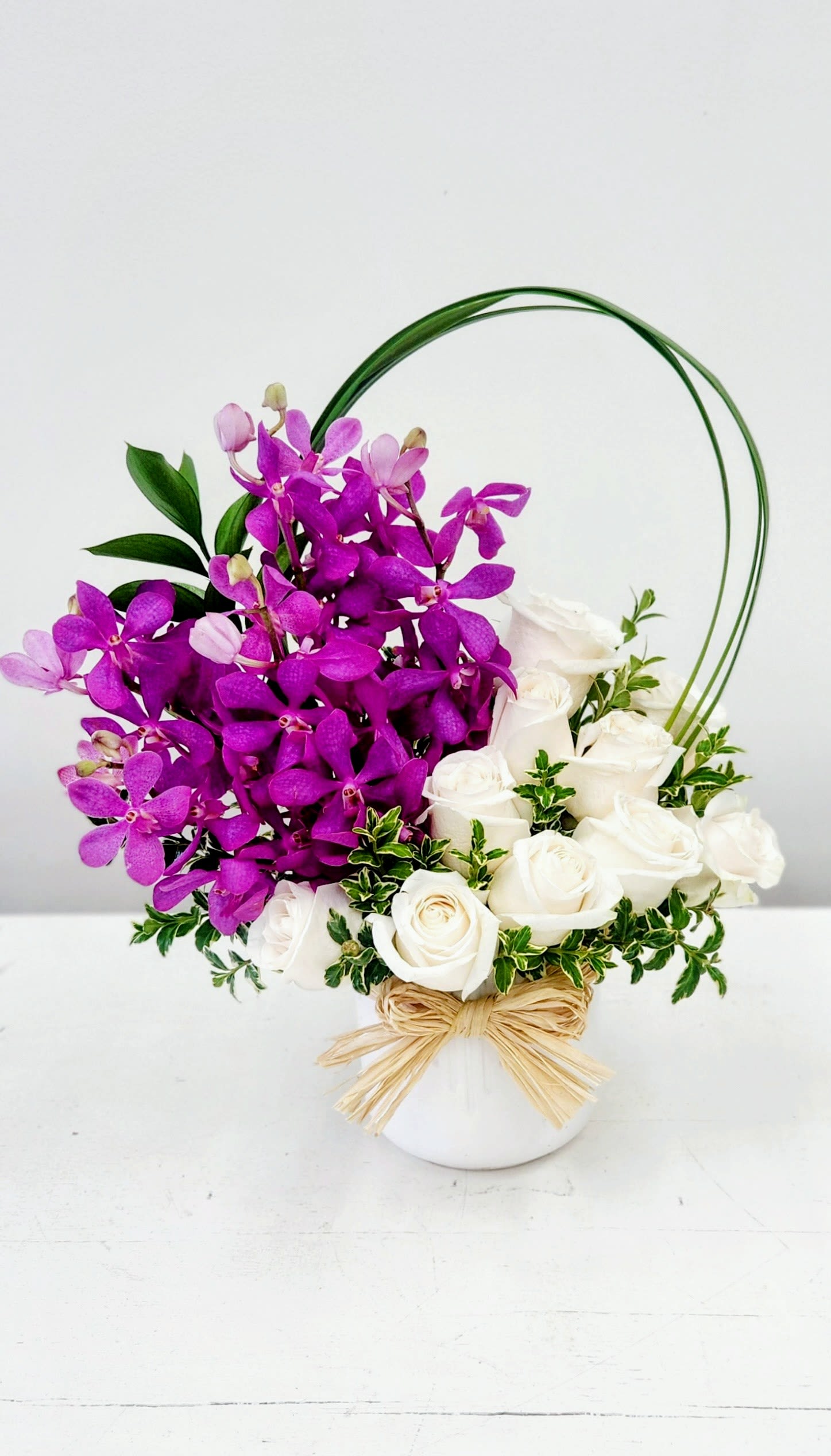 Orchids and White Roses - A dense cluster of vivid purple Dendrobium orchids paired with creamy white roses creates a striking contrast that feels both romantic and celebratory - perfect as a Mother's Day gift or to mark a birthday, retirement, or milestone. This flower arrangement's polished two-tone composition makes the recipient feel seen and celebrated, whether you're sending love and romance, a thoughtful get-well wish, or a simple "thinking of you" surprise.  