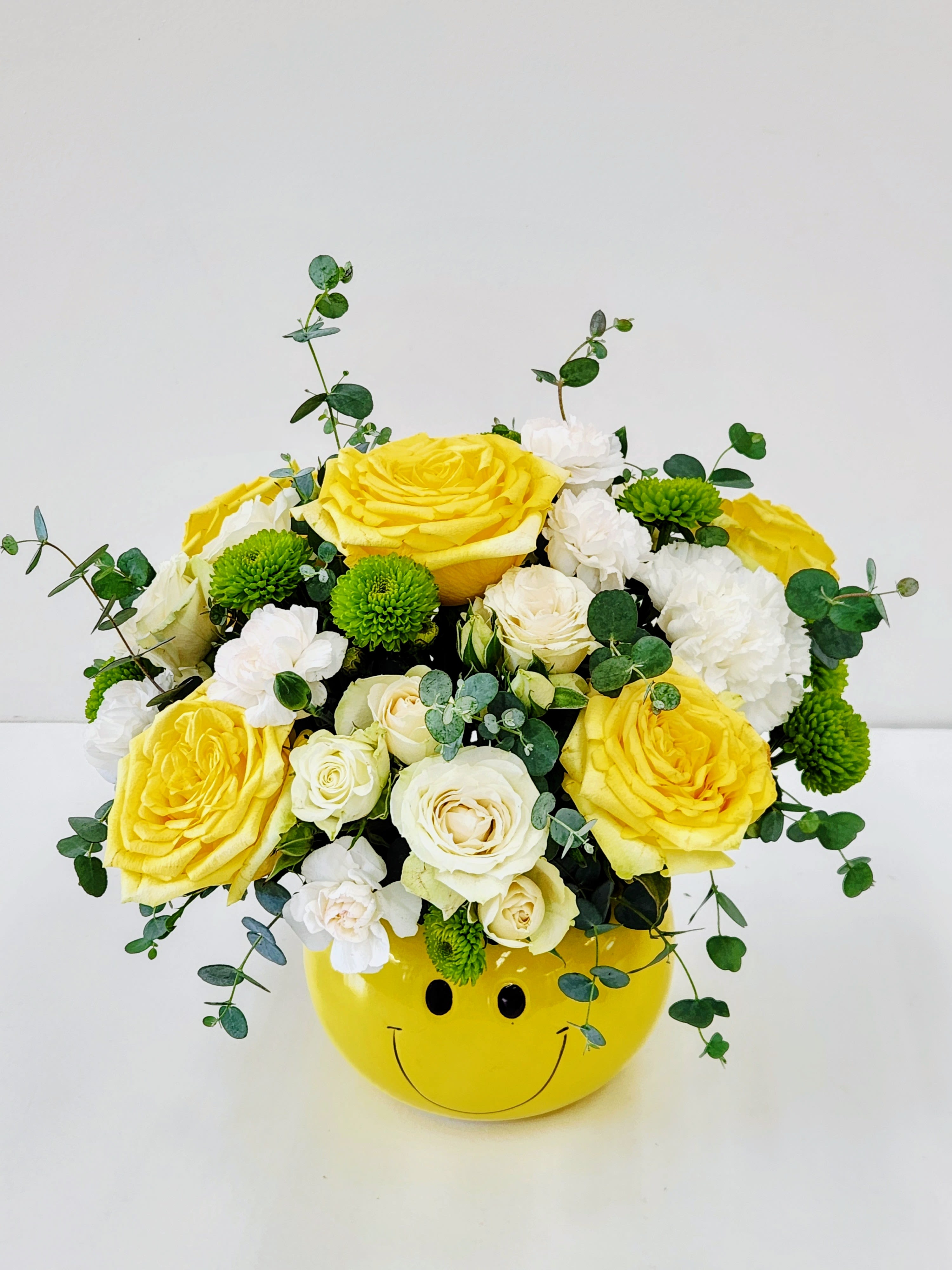 The Happy Face Bqt - Sunny yellow roses and crisp white blooms tucked into a playful yellow smiley-face ceramic container make this flower arrangement an instant mood lifter - perfect as a birthday bouquet, a cheerful congratulations gift, or a lighthearted just-because surprise. The recipient will feel genuinely seen and cheered by the bright, sunny palette and the arrangement's casual, upbeat personality.  