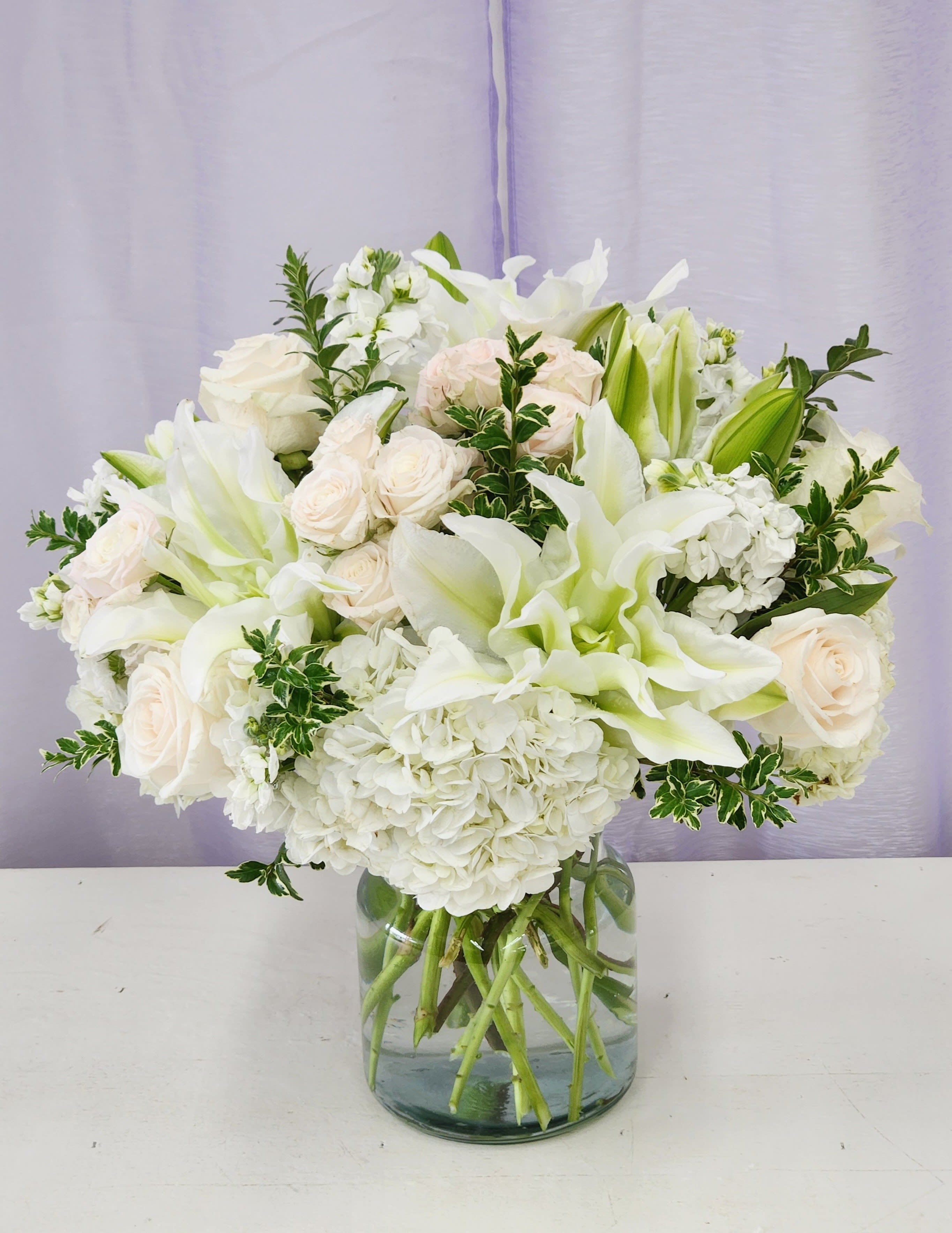 Pure Elegance - A pool of large white lilies and clustered white hydrangeas gives this bouquet an instantly serene, elegant presence-perfect as a wedding centerpiece, sympathy arrangement, or a refined anniversary bouquet. Soft blush and cream roses warm the predominantly white palette so the person you're thinking of feels comforted and celebrated rather than overwhelmed; bright green lily buds and variegated foliage add fresh contrast and visual lift.   